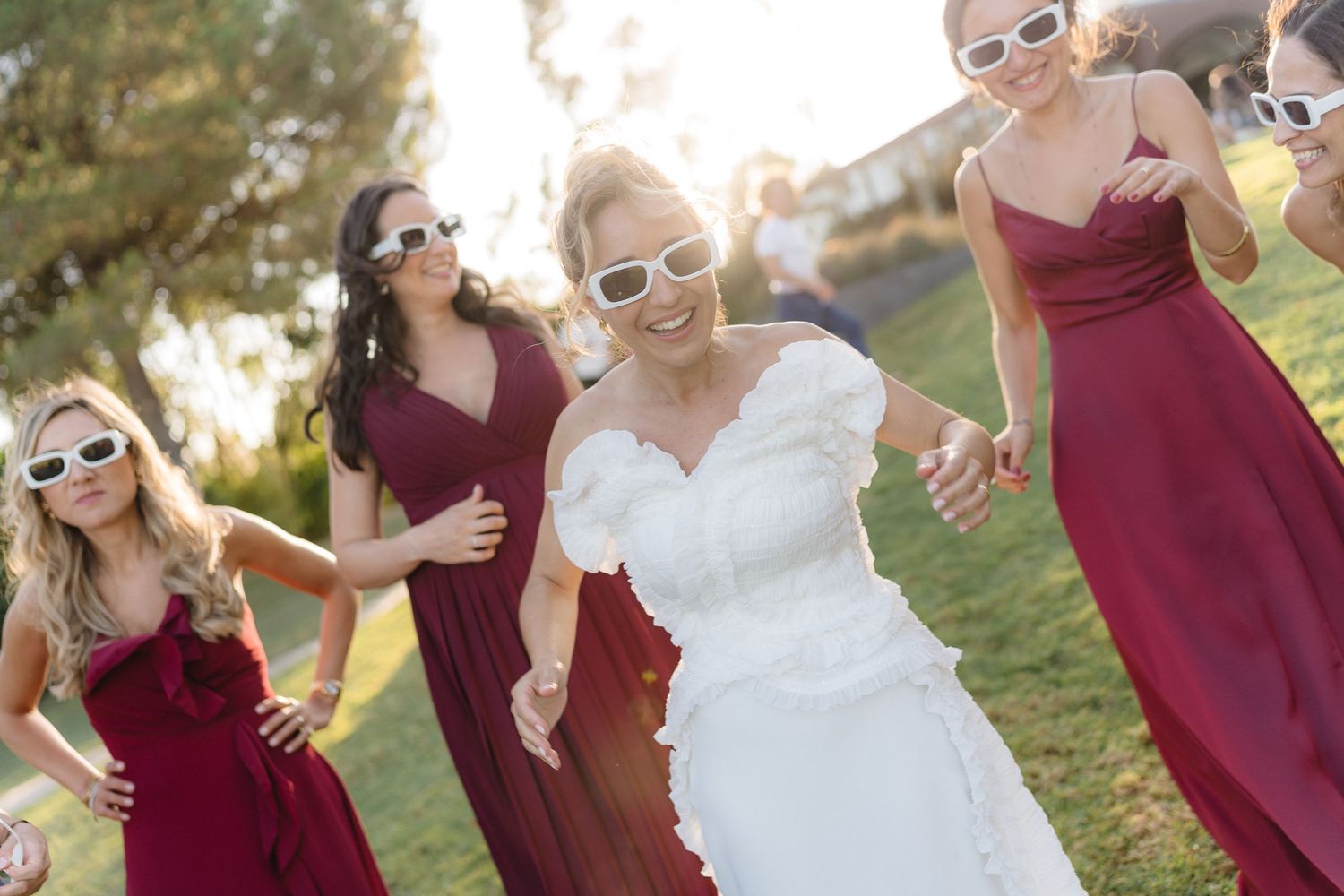 Wedding party in burgundy dresses and white sunglasses poses playfully outdoors.