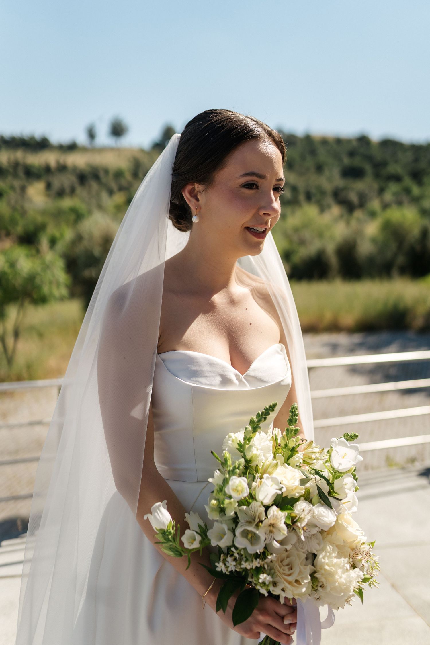 A bride in a classic strapless gown holds a white bouquet against a sunny outdoor backdrop.