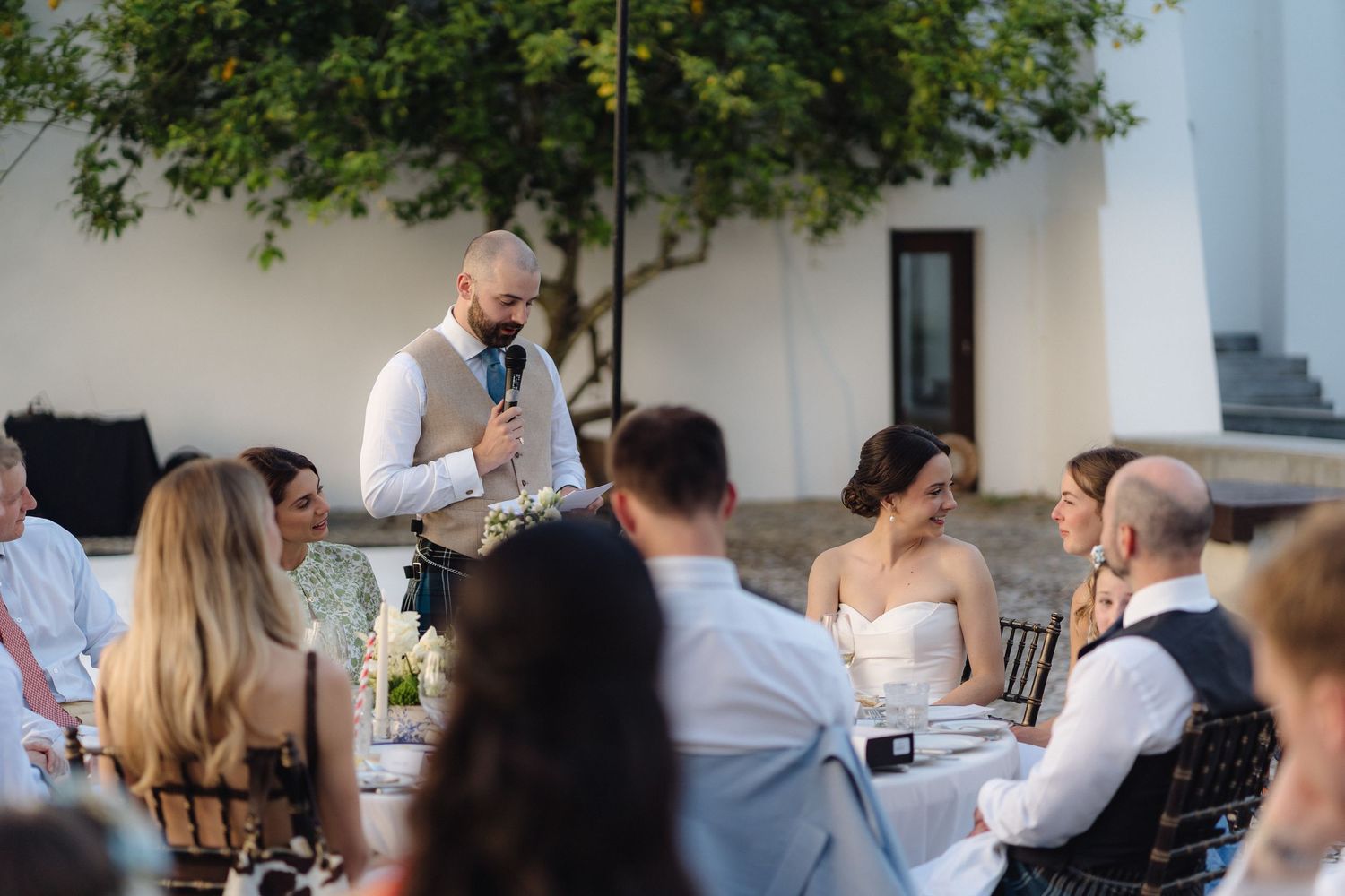 A speaker addresses wedding guests during an outdoor reception under trees.