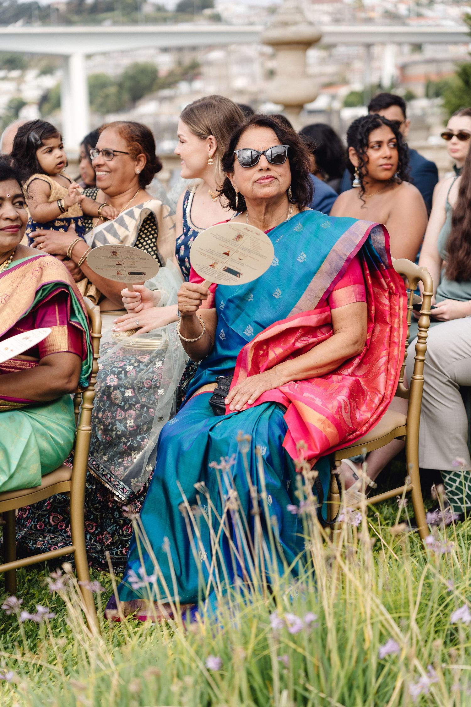 Wedding guests seated outdoors in colorful attire watching ceremony in garden setting.