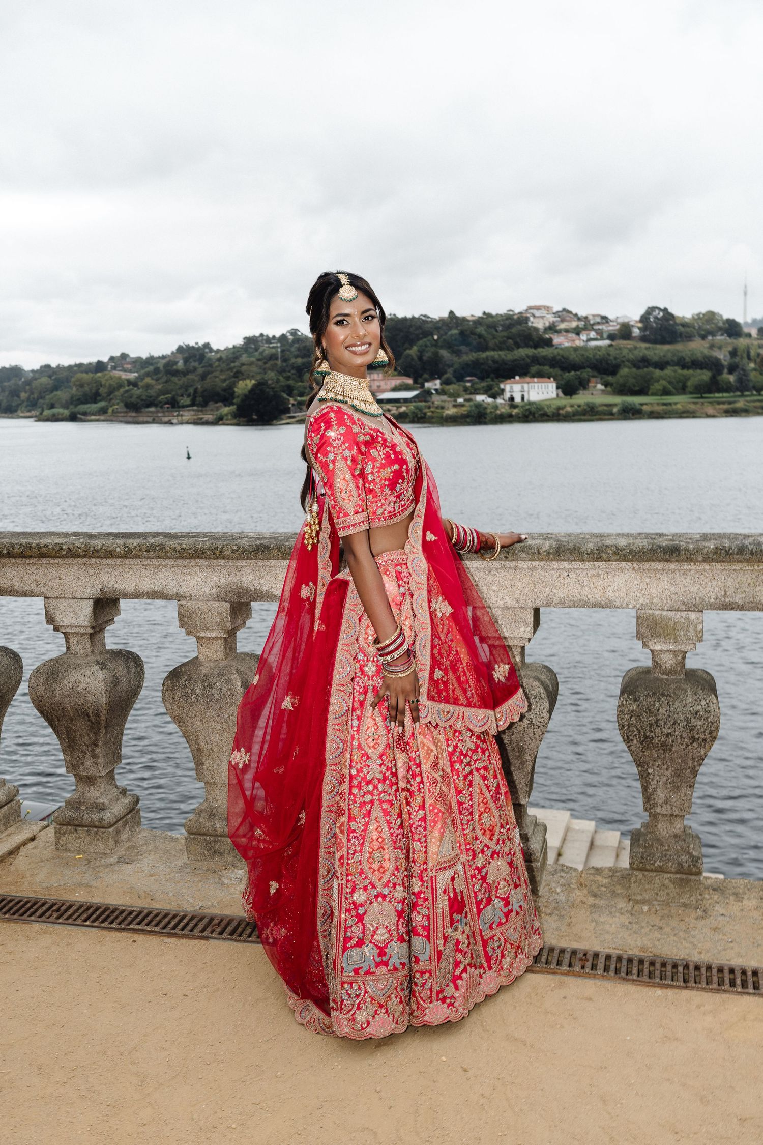 Traditional red bridal lehenga photographed against waterfront stone architecture.