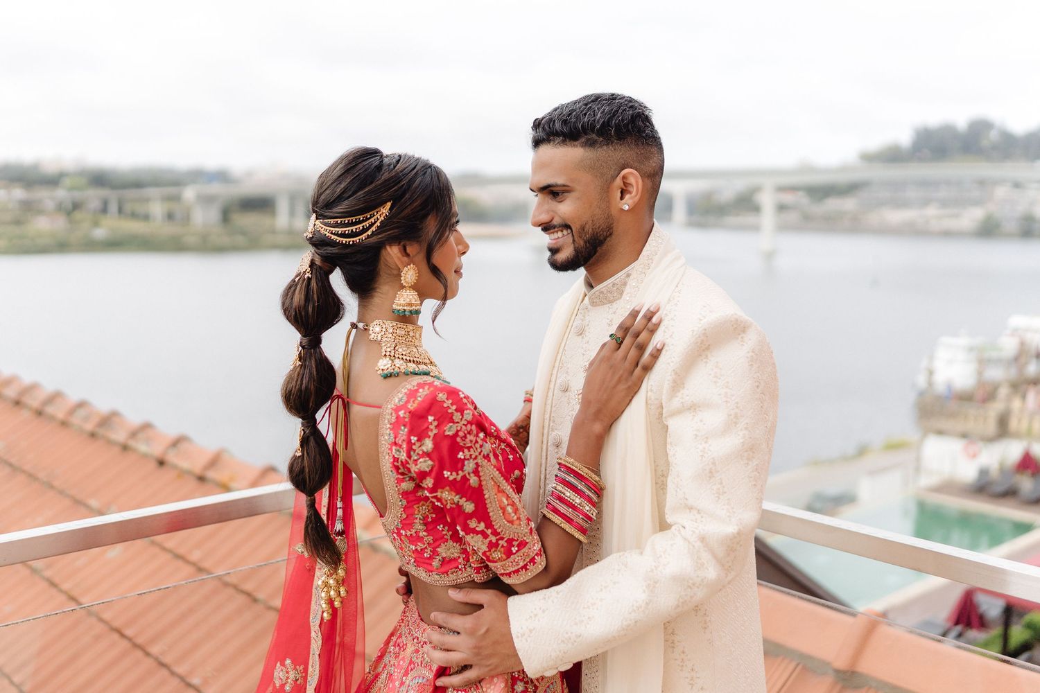 Romantic couple in traditional Indian wedding attire posing by waterfront balcony.