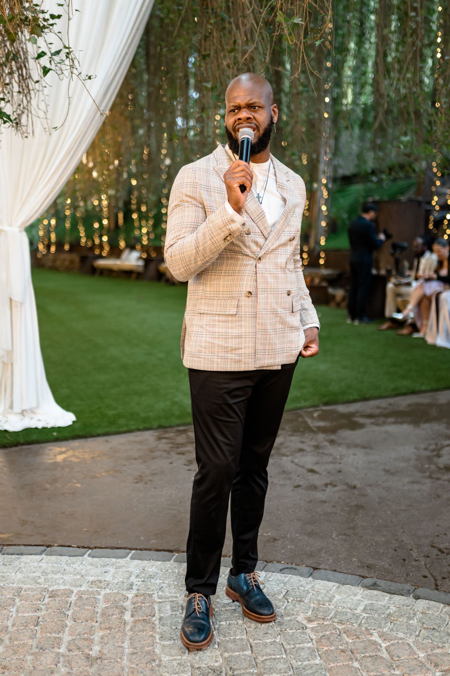Wedding guest in a cream checkered suit jacket stands on cobblestone pavement during an outdoor celebration.