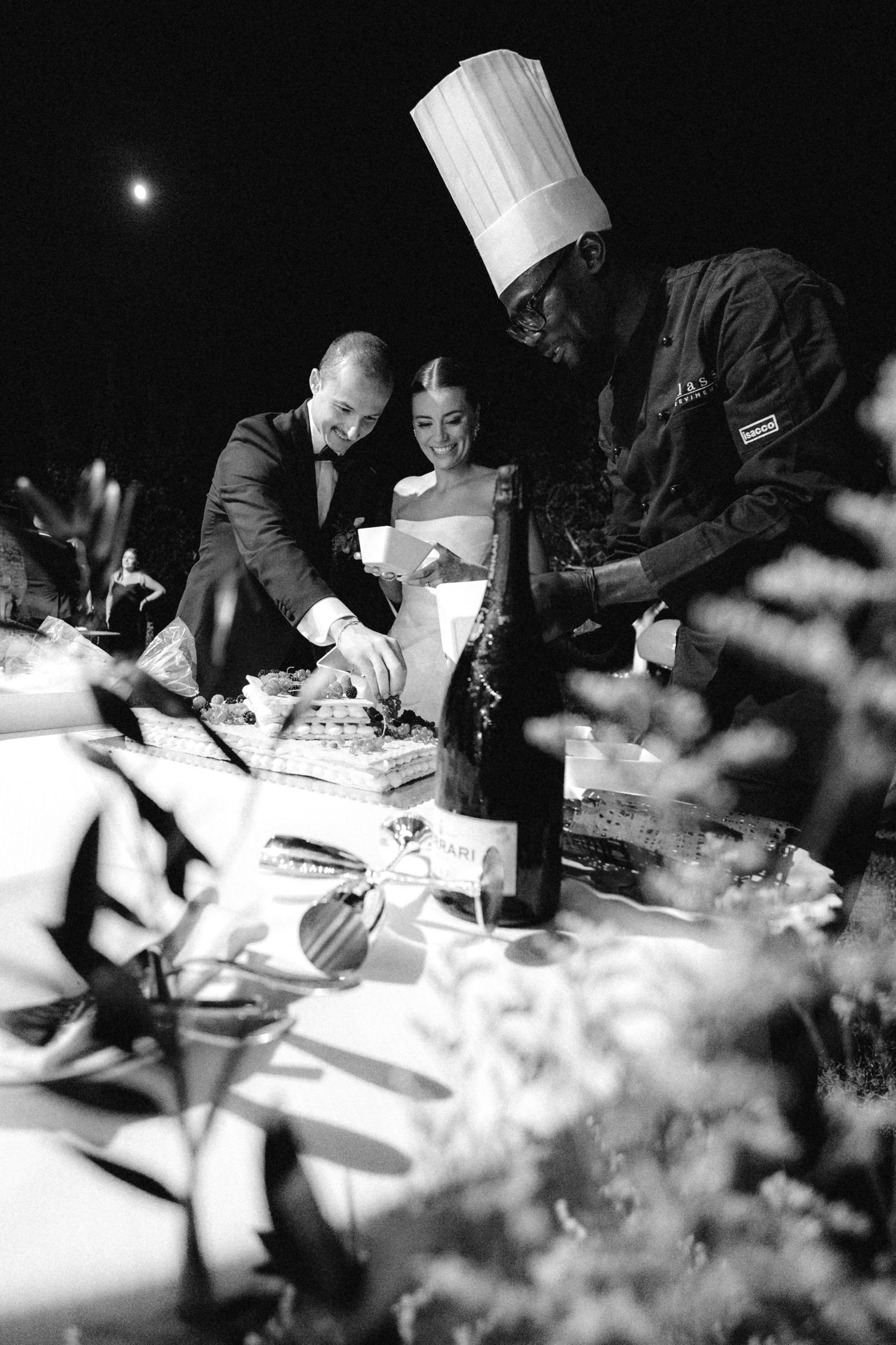 Black and white photo of a bride and groom cutting cake assisted by a chef at an outdoor wedding reception.