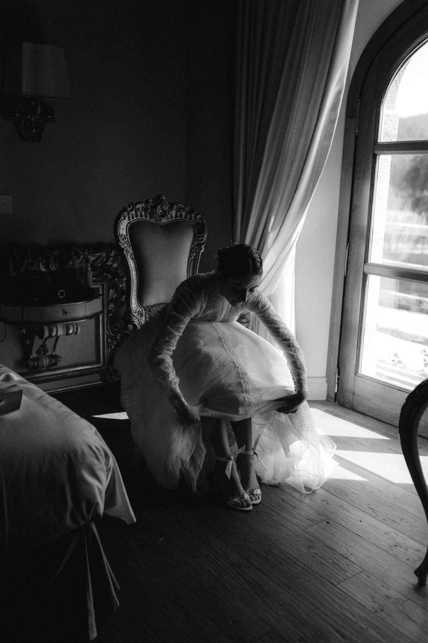 Bride in white wedding gown adjusting her dress near a sunlit arched window in an elegant room.
