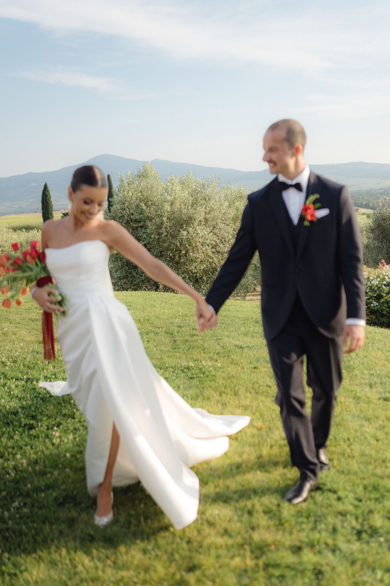 Newlyweds holding hands walking on green lawn with Tuscan hills and olive trees behind them.