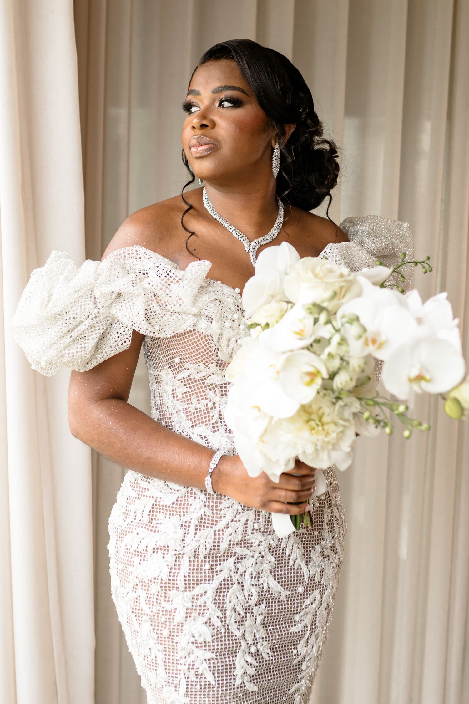 A bride in an intricately detailed lace wedding dress holds a white orchid bouquet.