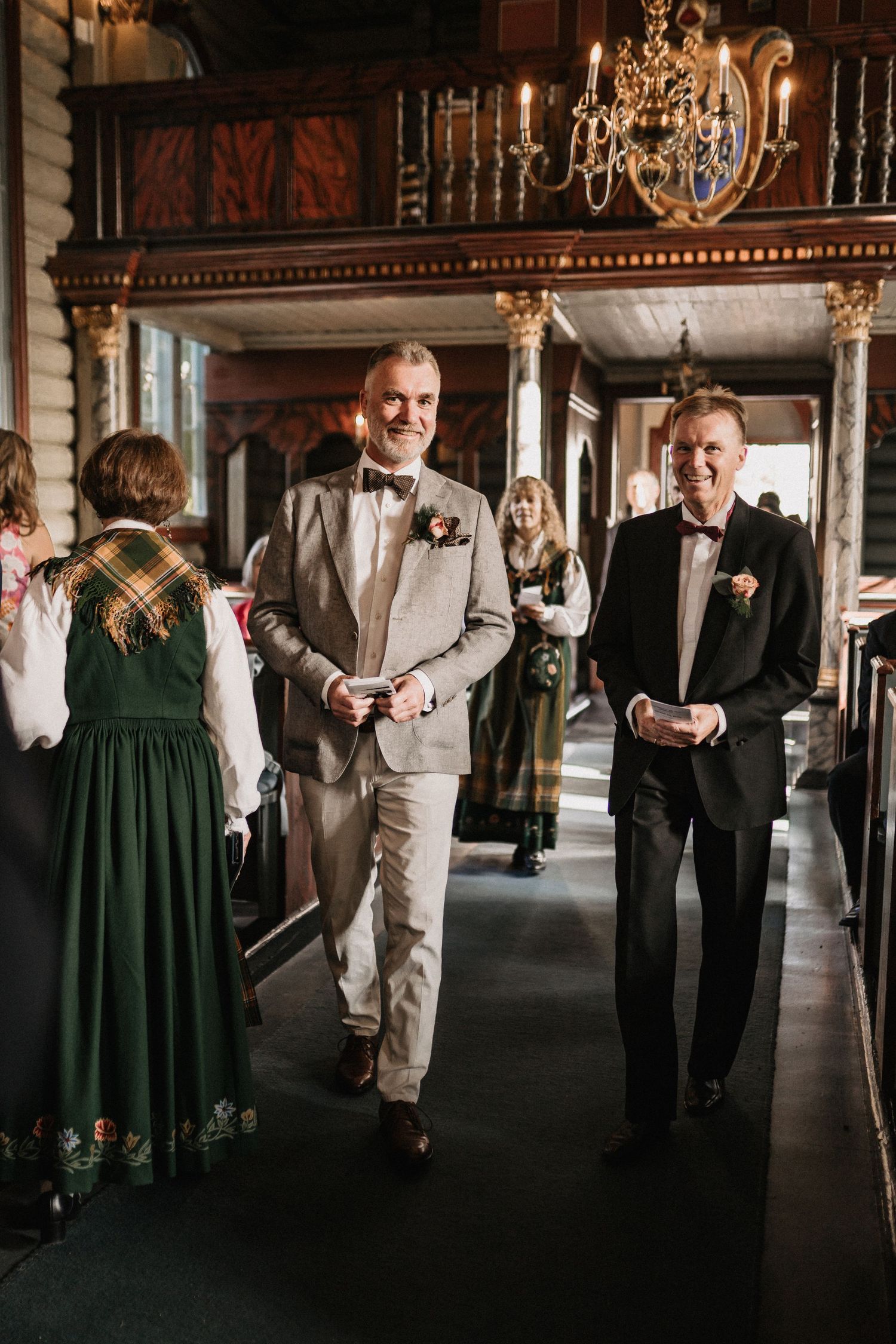 Two elegantly dressed guests walk down an ornate hallway with chandelier and traditional decor at a formal event.