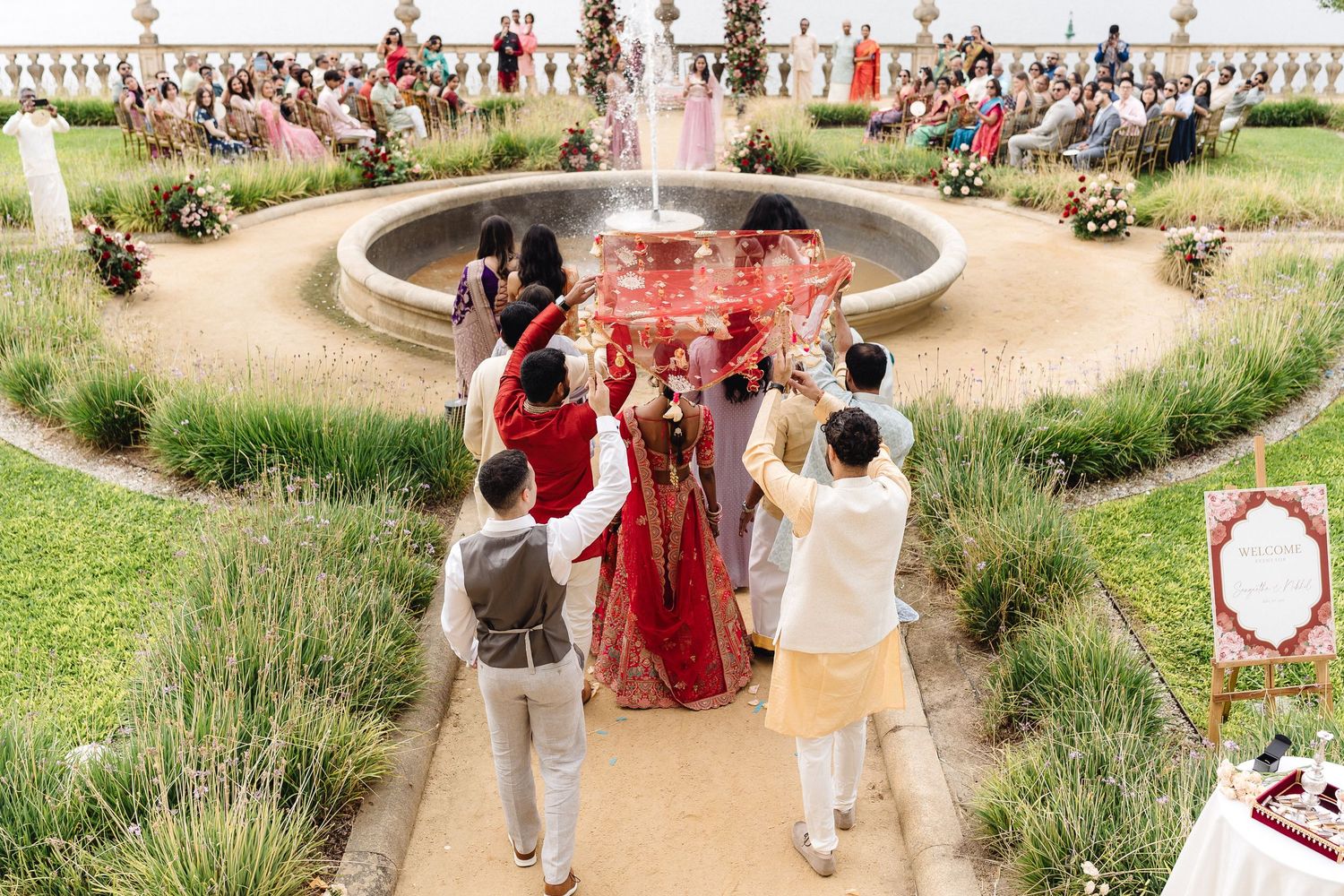 A traditional Indian wedding ceremony takes place outdoors with guests gathered around a floral-decorated mandap.