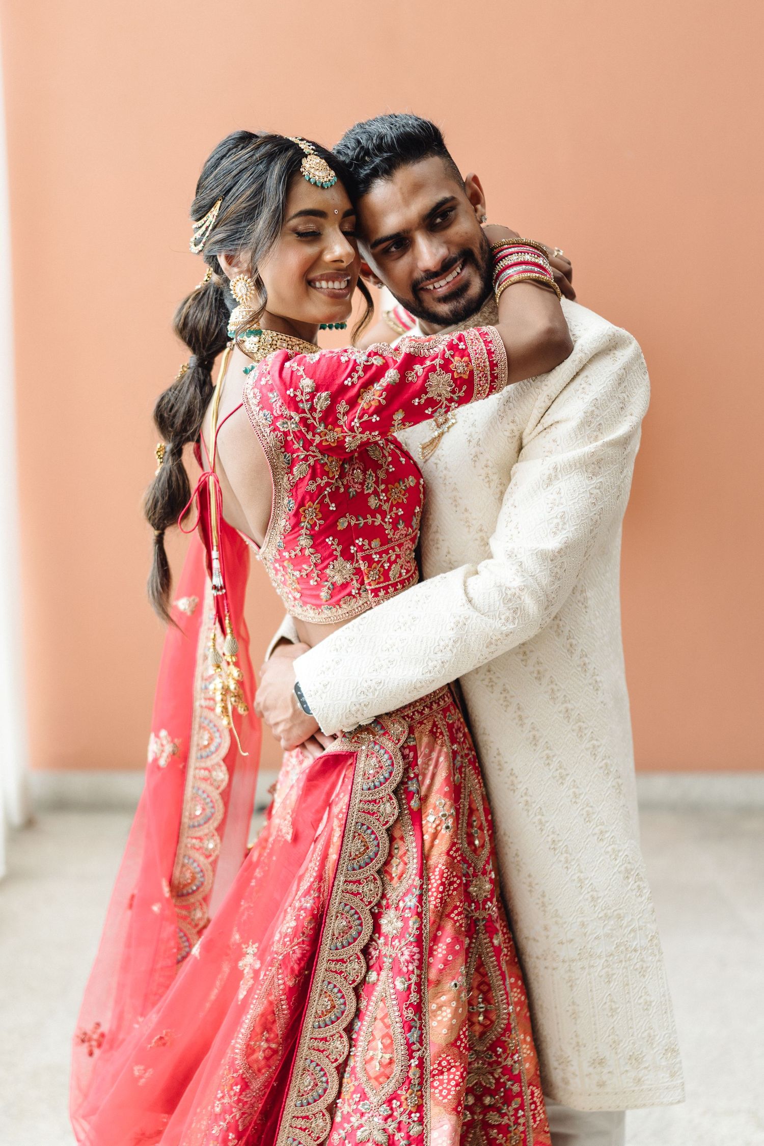 Romantic couple in traditional Indian wedding attire embracing against peach-colored wall.