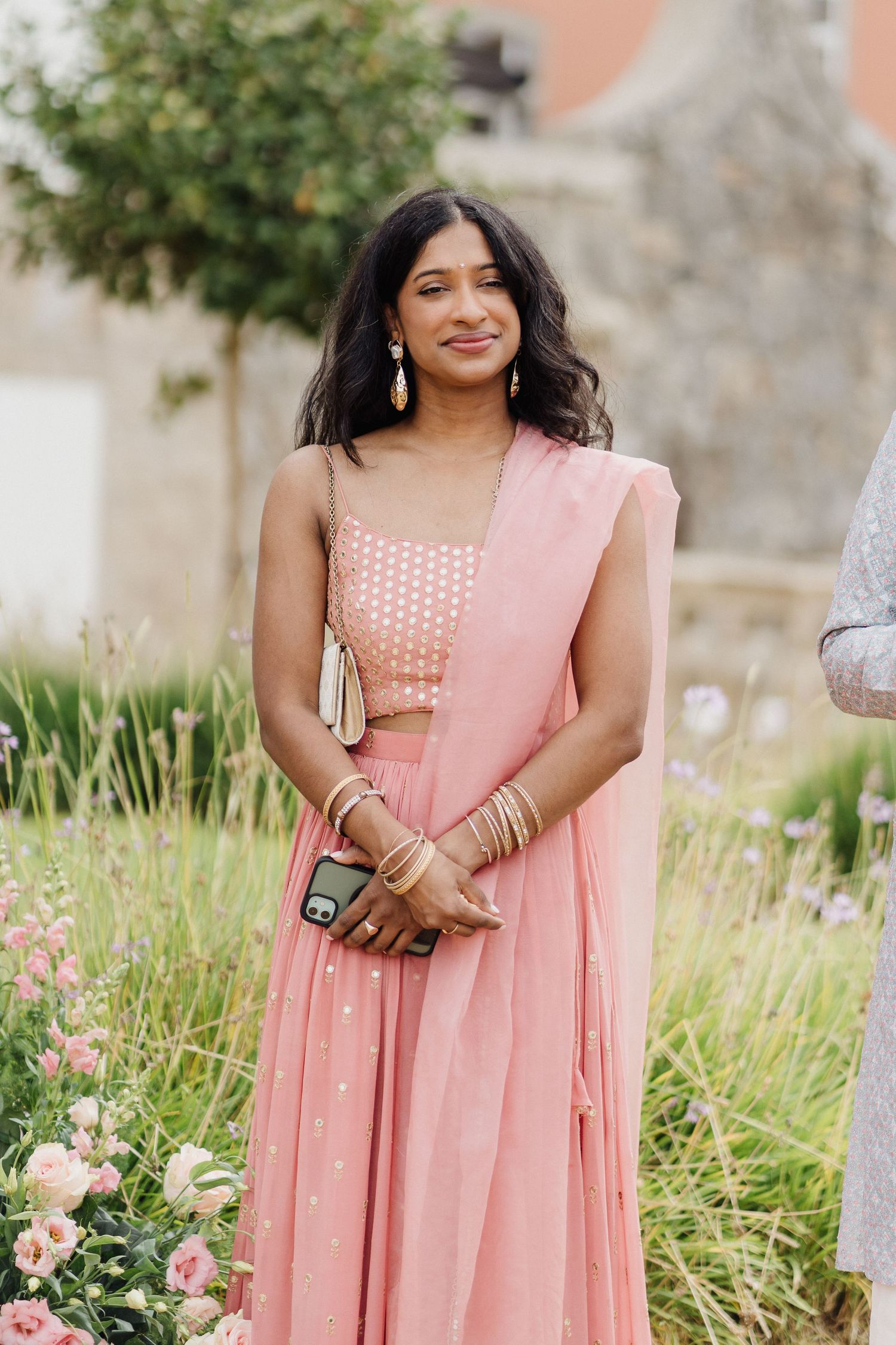 Woman in a soft pink traditional Indian dress standing in a garden with blooming flowers.