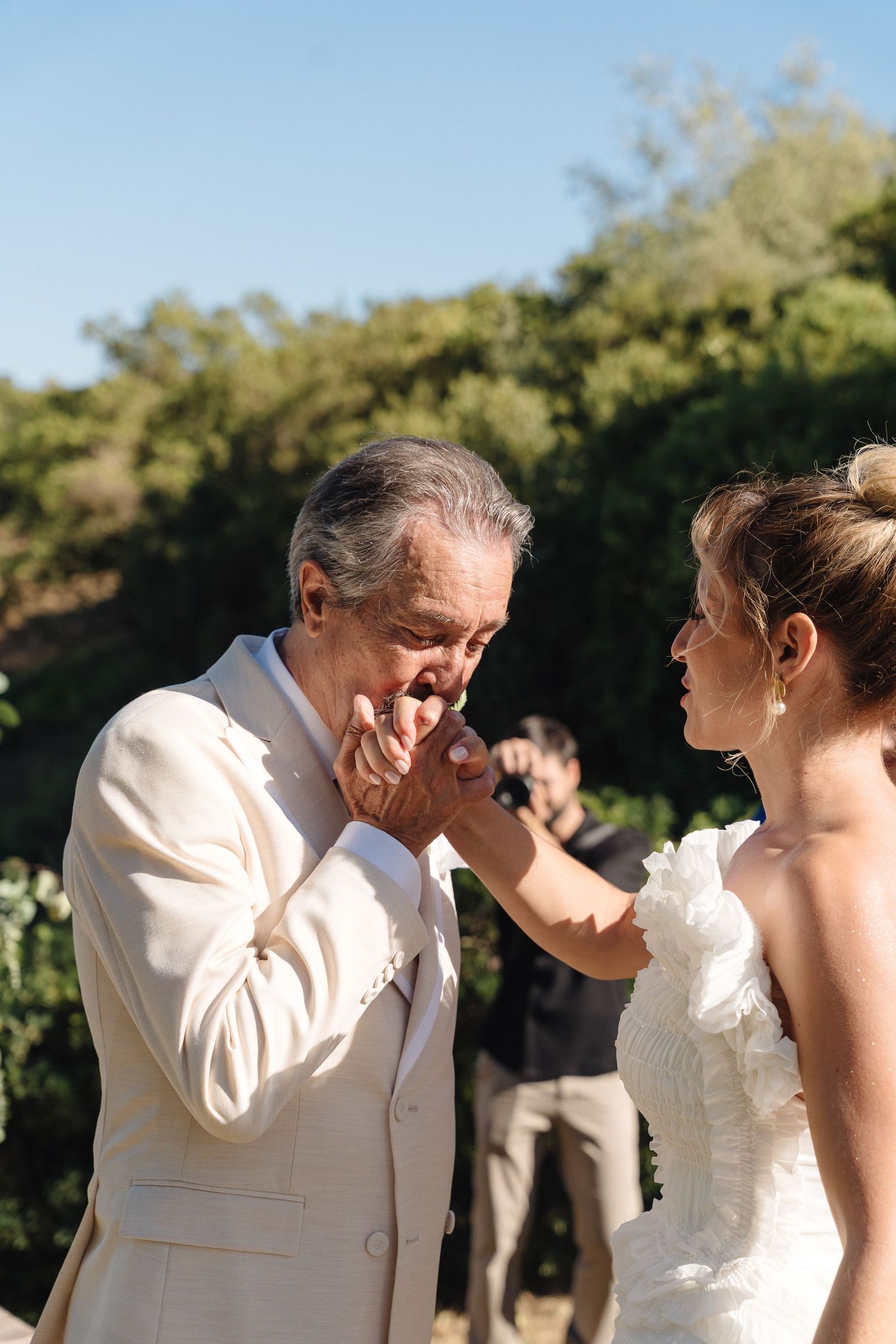 An emotional moment shared between a bride in white and older man in cream suit during an outdoor wedding ceremony.