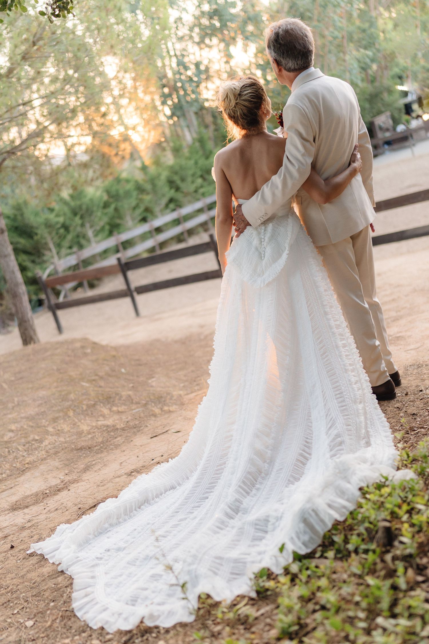 A couple walks together on a garden path during sunset, with the bride's lace wedding dress trailing behind them.