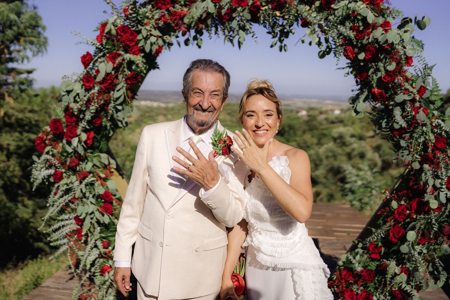 Wedding couple laughing under a floral arch decorated with red roses and greenery.