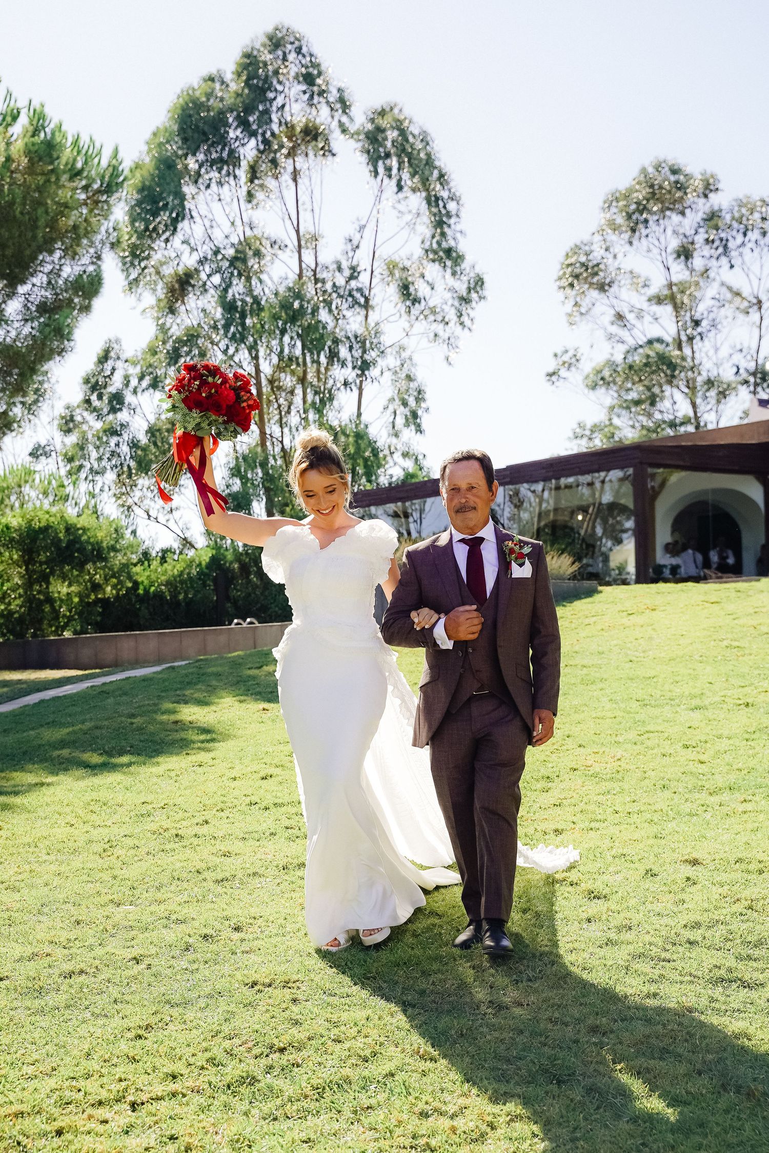 A couple in wedding attire walks on grass with red bouquet raised triumphantly.
