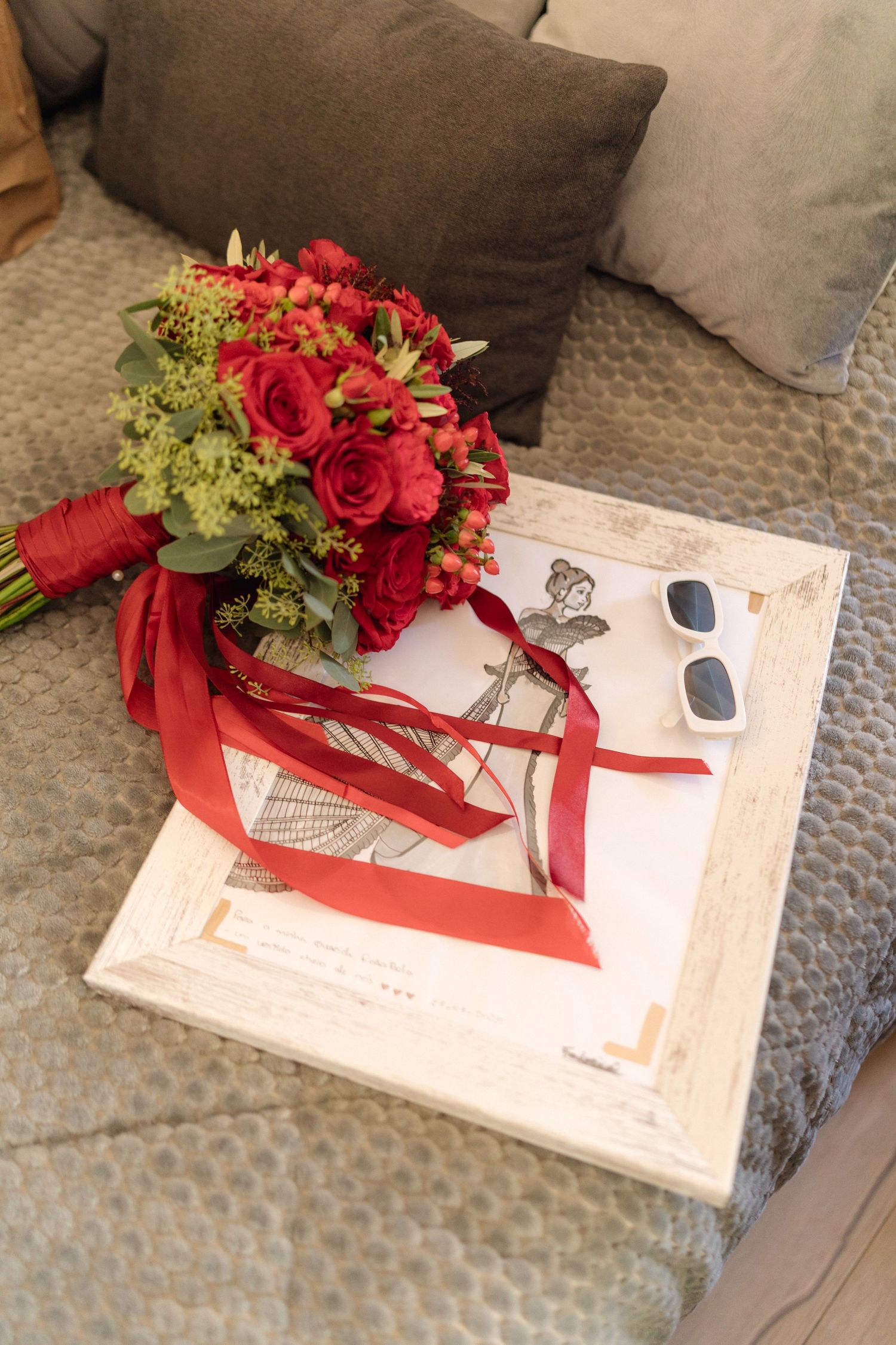 Red rose bouquet with sunglasses and ribbons arranged on grey couch with decorative frame.