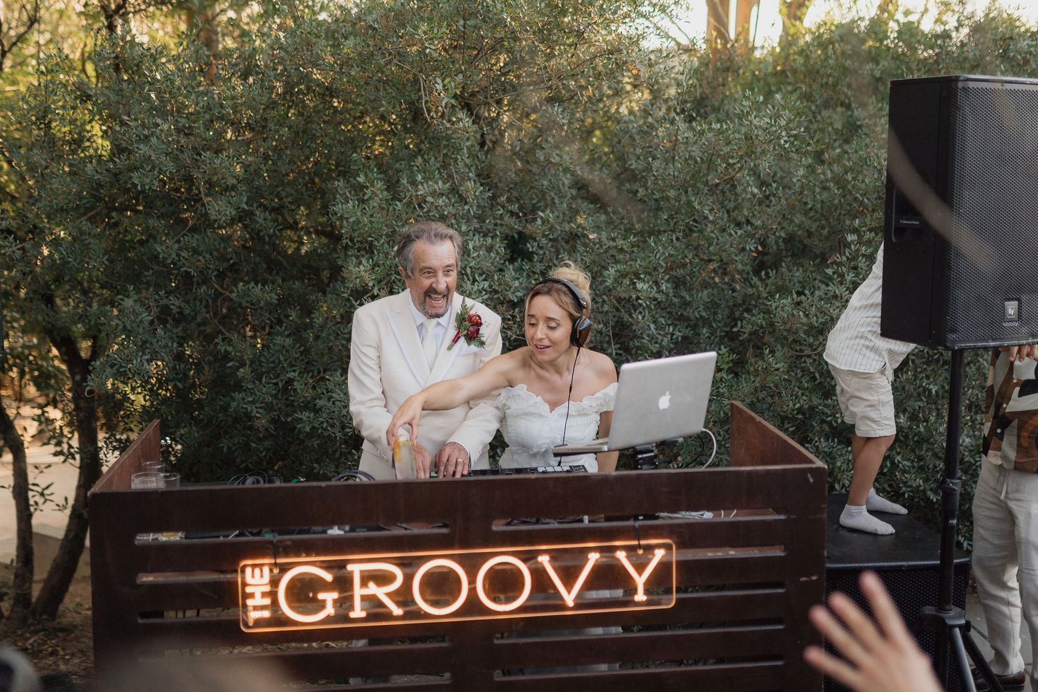 DJs entertaining guests at outdoor reception with illuminated 'Groovy' sign display.