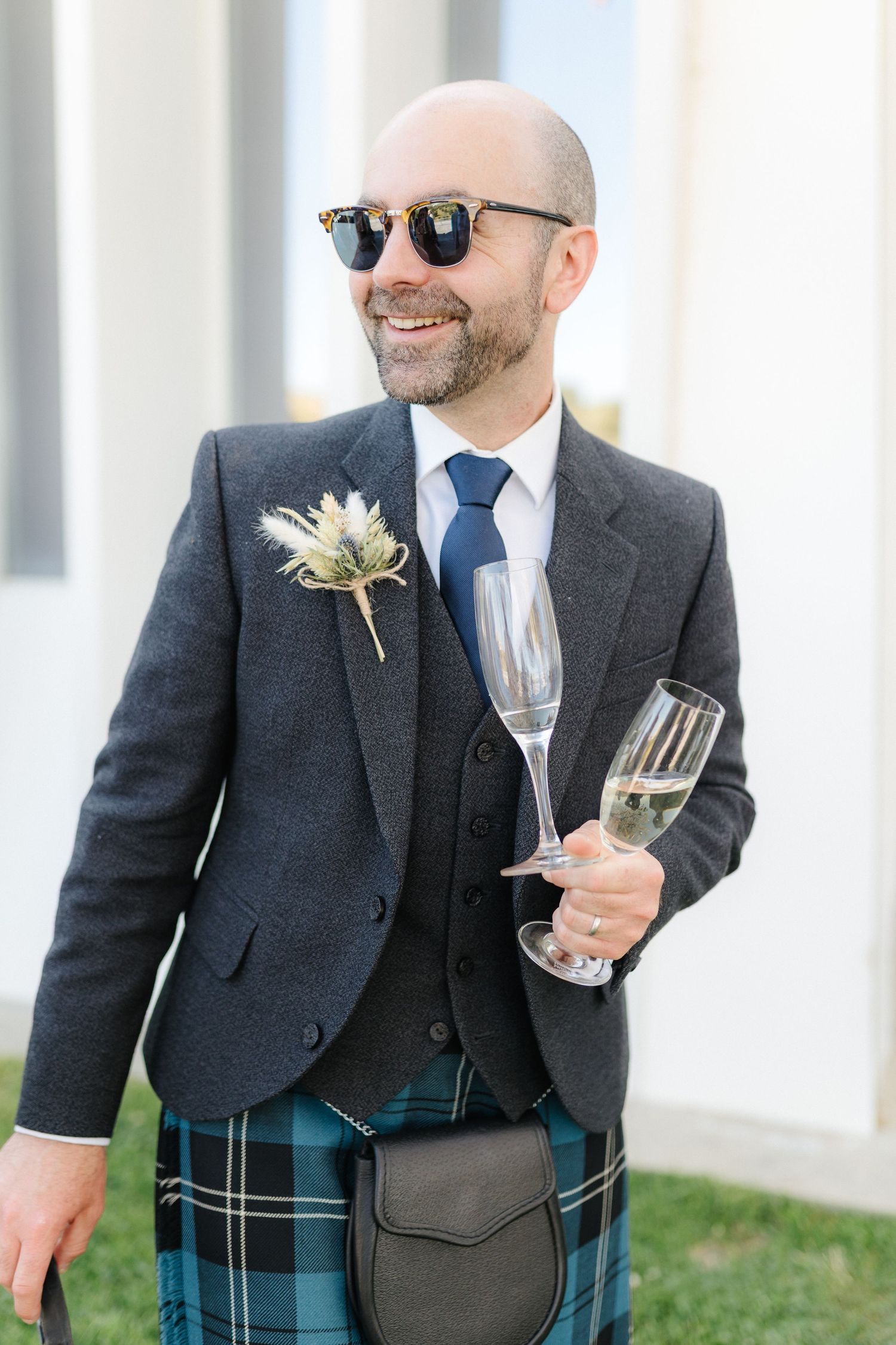 Groomsmen in dark suits and kilts holding wine glasses at outdoor reception.