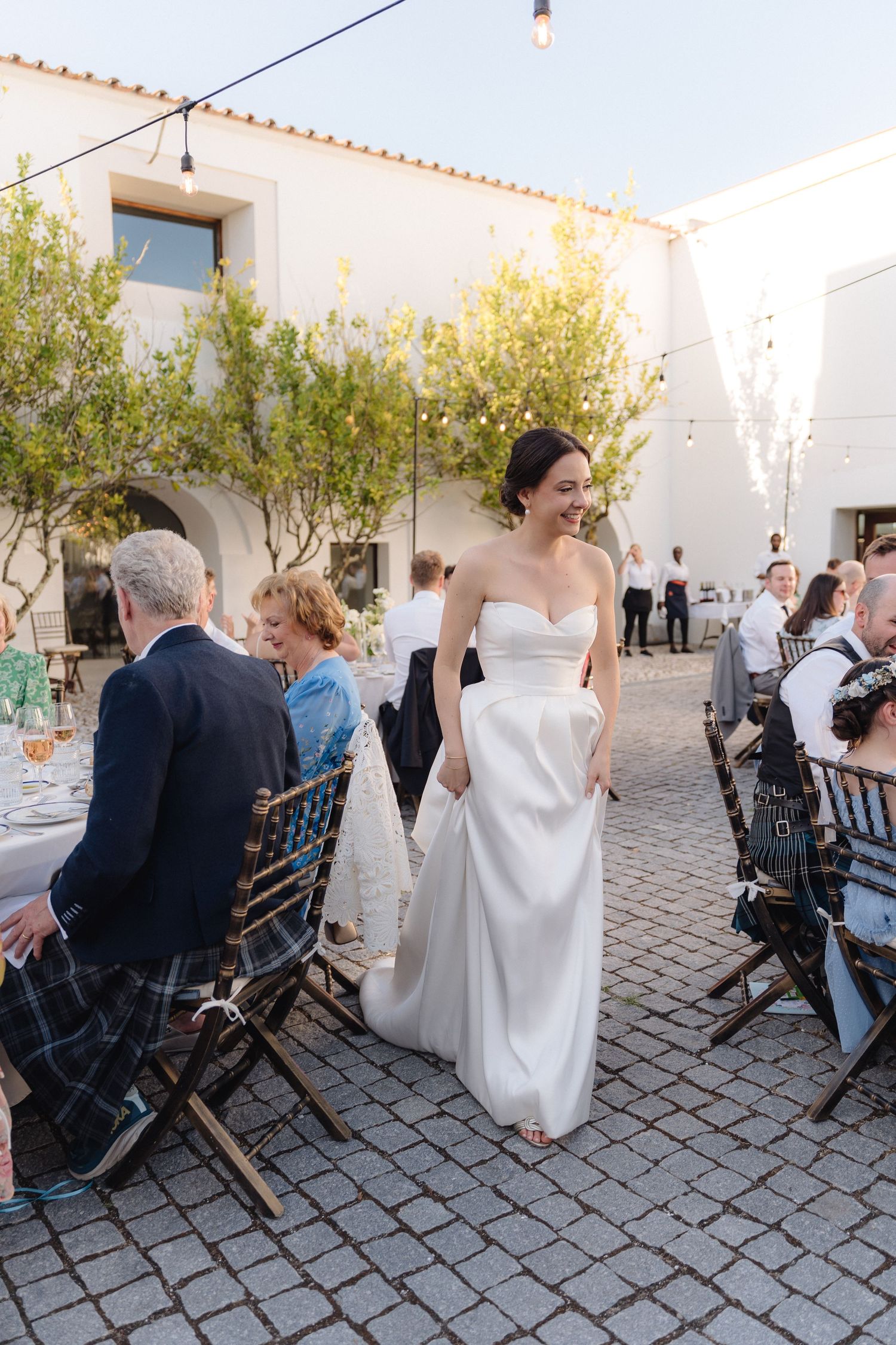 Wedding reception setup with wooden chairs on cobblestone patio surrounded by white buildings.