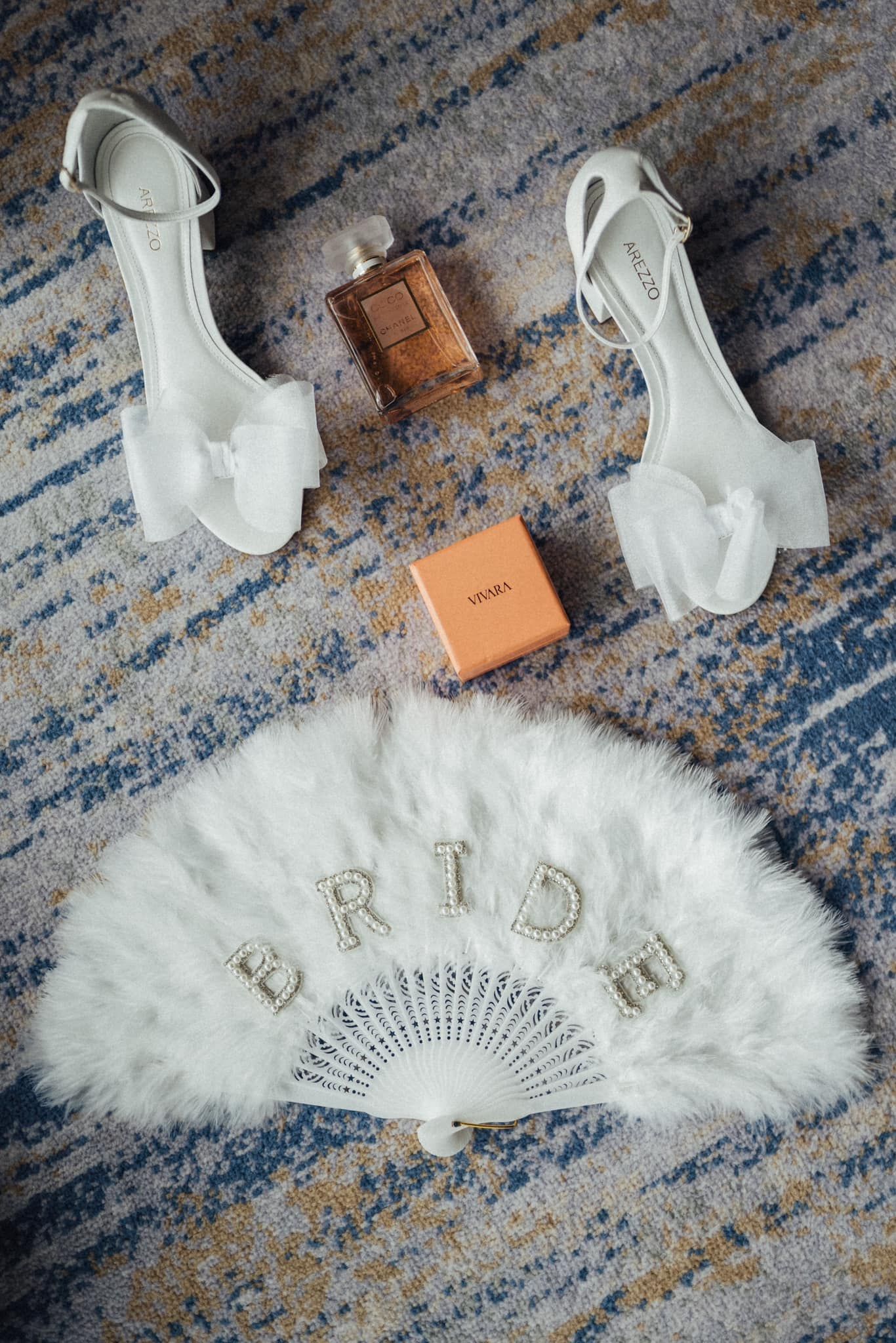 White bridal accessories including heels, fan, and perfume arranged on a blue patterned carpet.