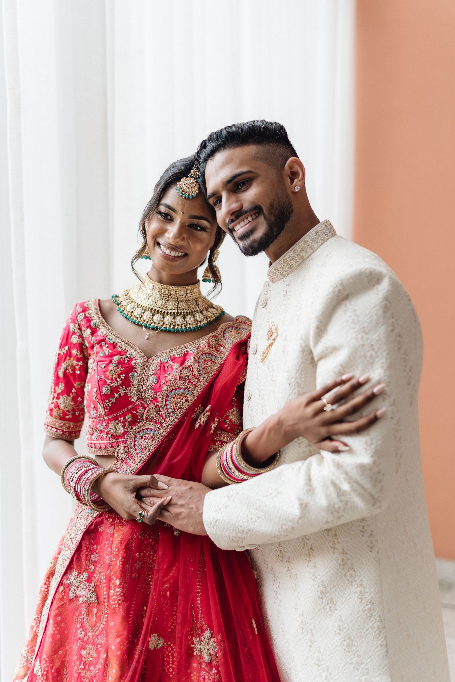 Wedding couple in traditional red lehenga and cream sherwani against white background.