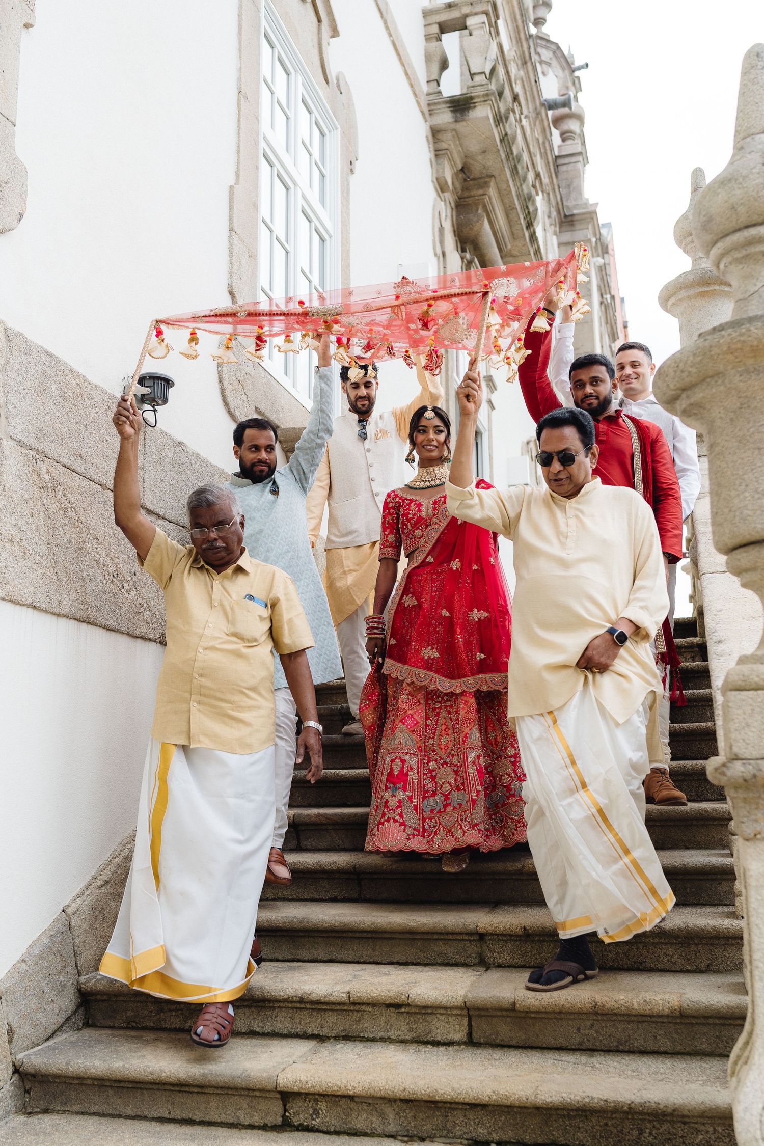 Wedding party poses joyfully on stone steps with traditional Indian attire and red fabric canopy.