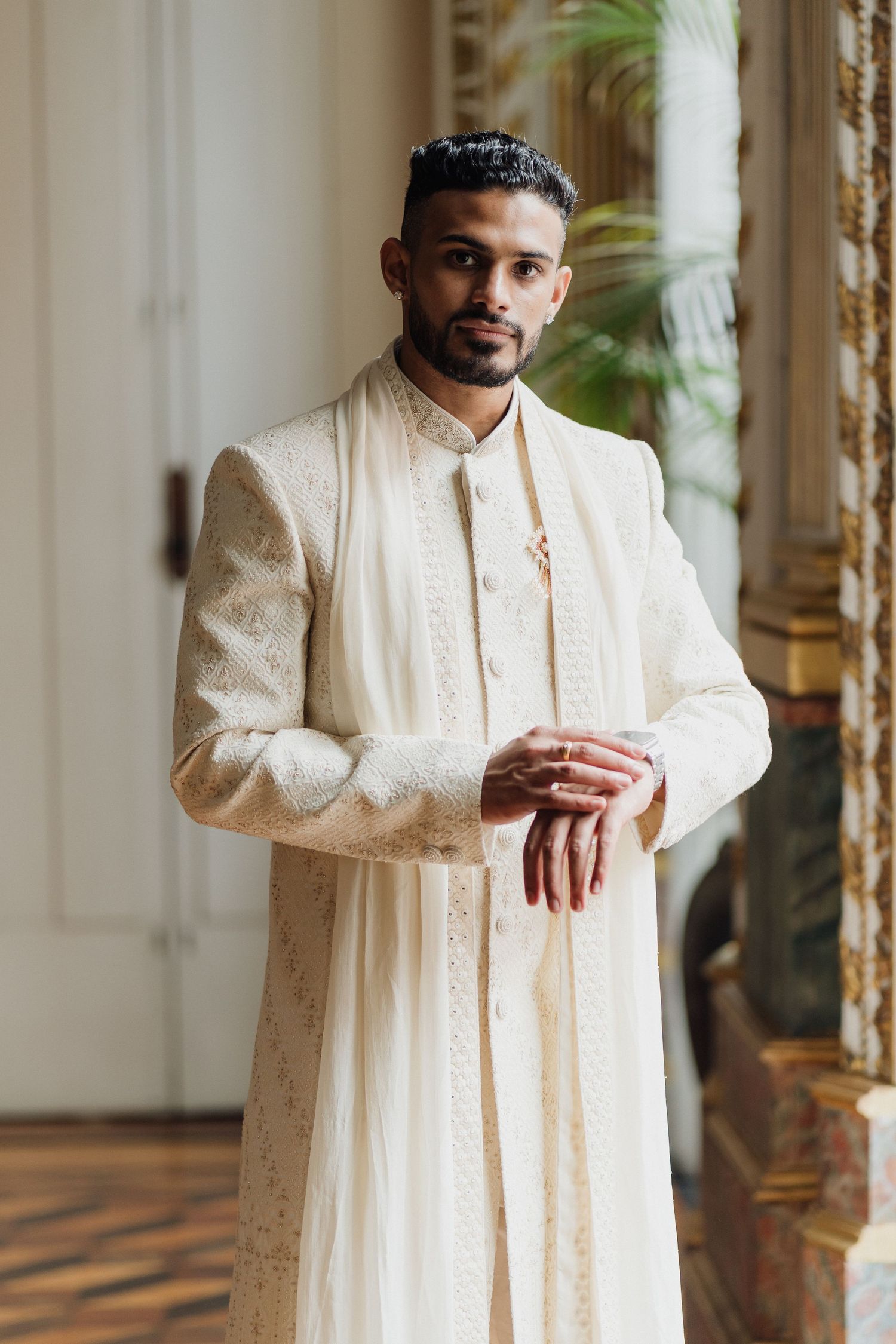 Groom wearing traditional cream sherwani with detailed embroidery in elegant indoor setting.