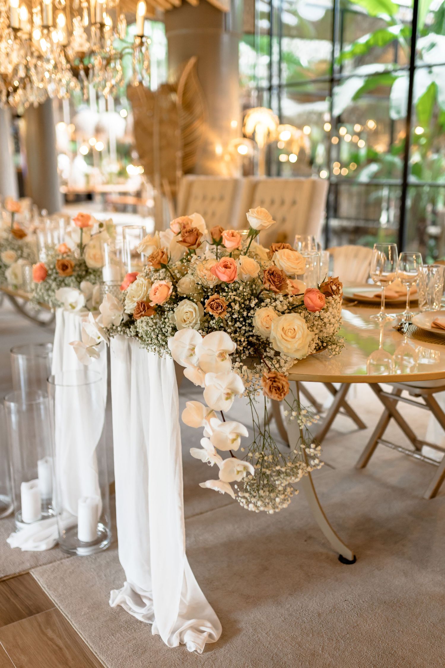 Elegant wedding table centerpieces featuring peach and white flowers with baby's breath in crystal vases under chandeliers.