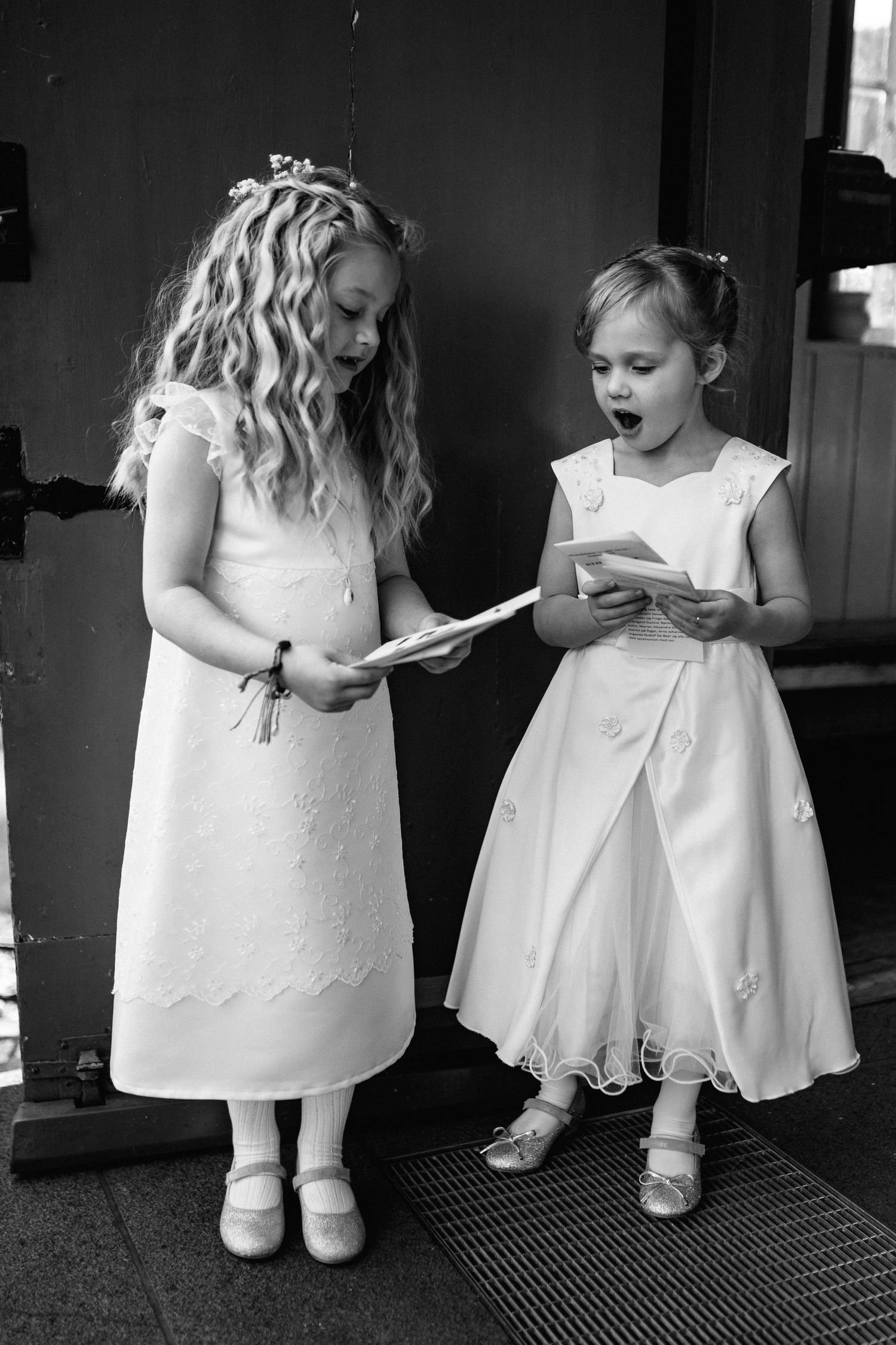 Young flower girls in white dresses sitting together on dark carpet.