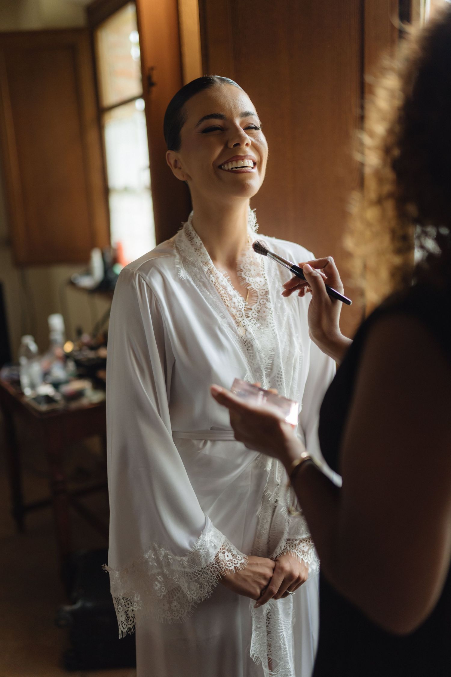 A smiling bride in a white lace robe gets makeup applied by a makeup artist during wedding preparations.