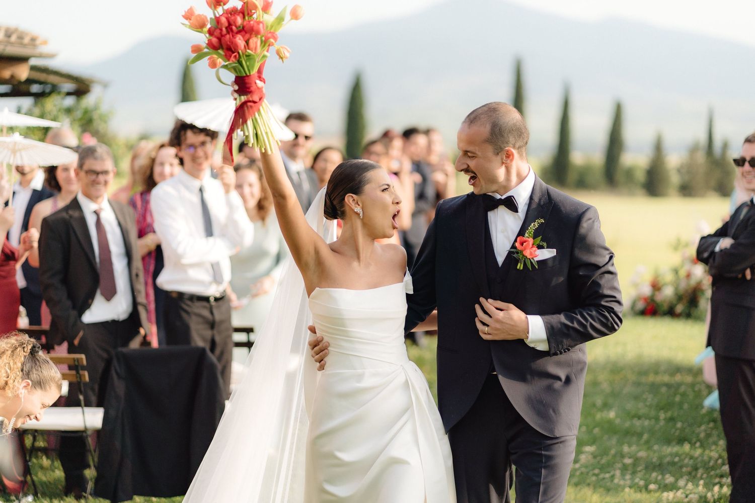 Bride raises bouquet of red tulips while walking with groom after outdoor wedding ceremony in Tuscany.