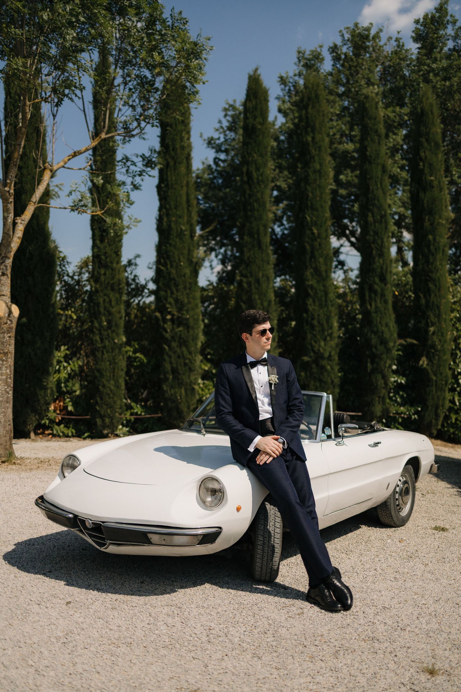 Stylish groom in navy tuxedo leaning on vintage white Alfa Romeo convertible among cypress trees.