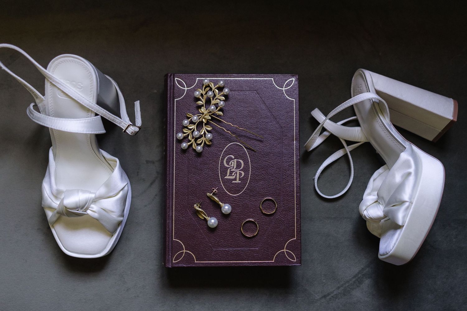 Wedding accessories including white shoes and jewelry arranged on dark surface.