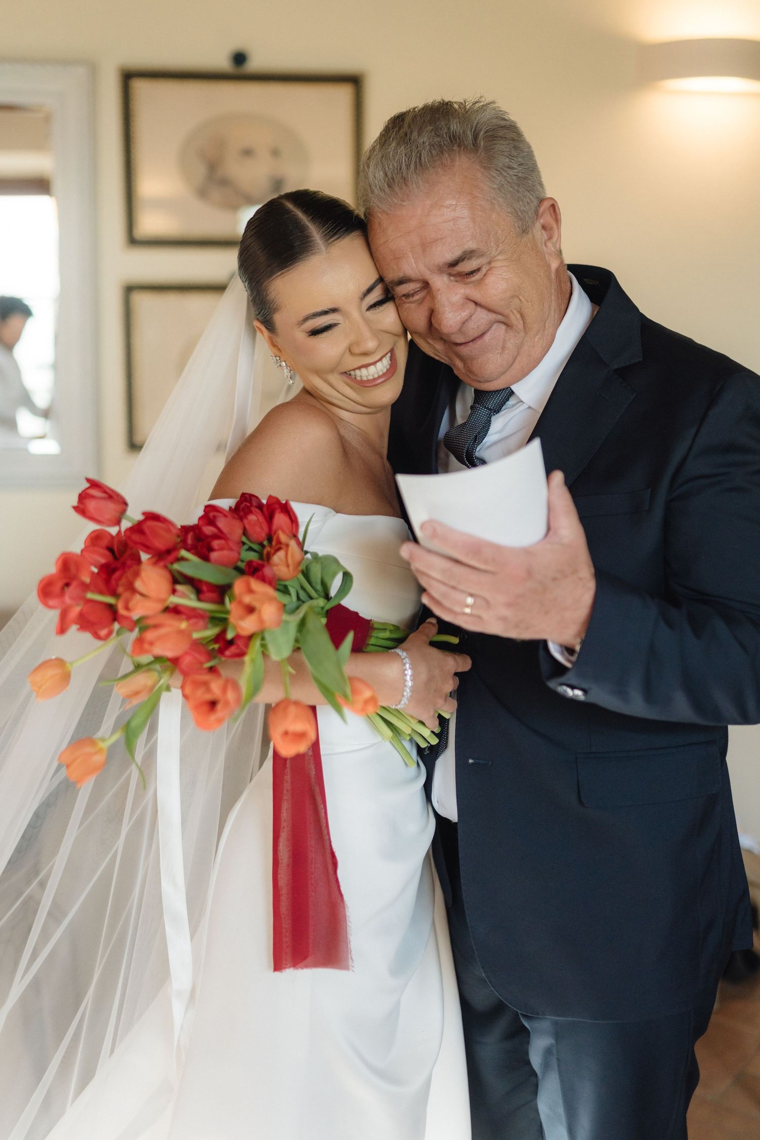 Bride in white gown holding red tulip bouquet shares joyful hug with older man in navy suit on wedding day.