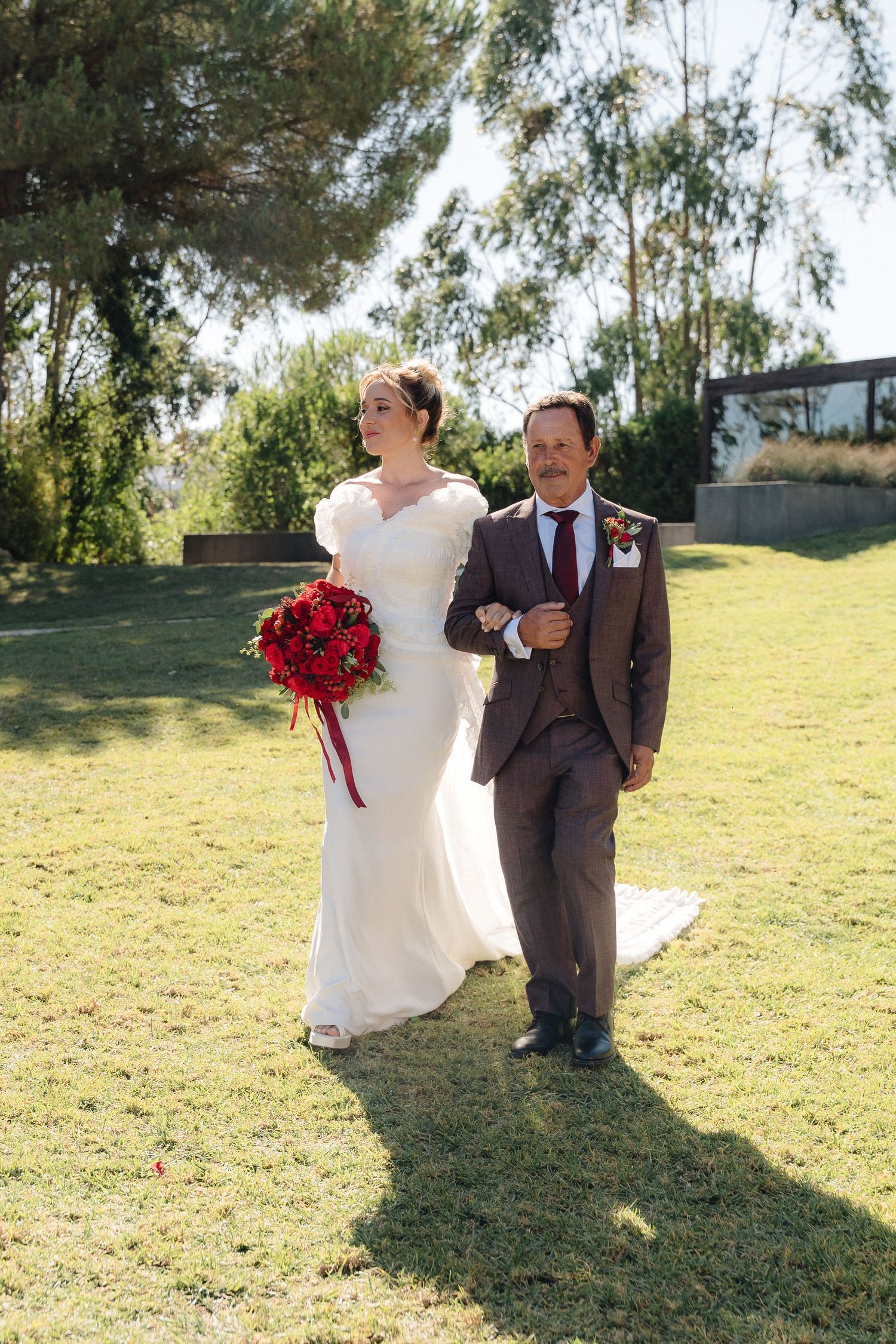 A couple in wedding attire walks arm-in-arm across a sunny lawn with red bouquet.