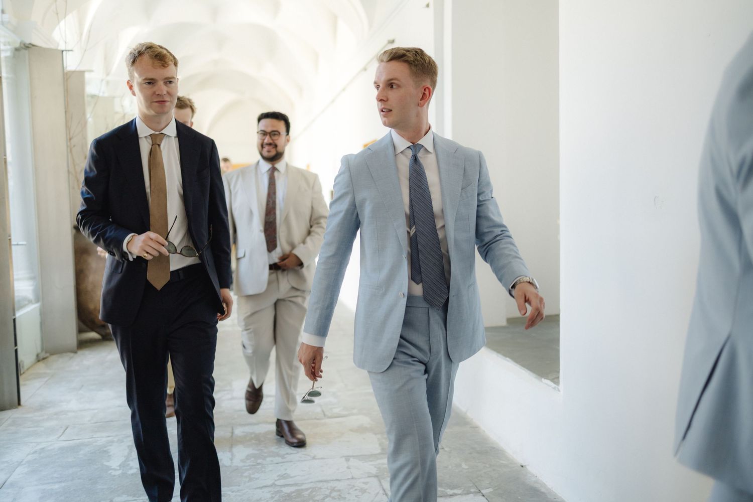 Men in formal suits walking through white corridor with natural lighting.