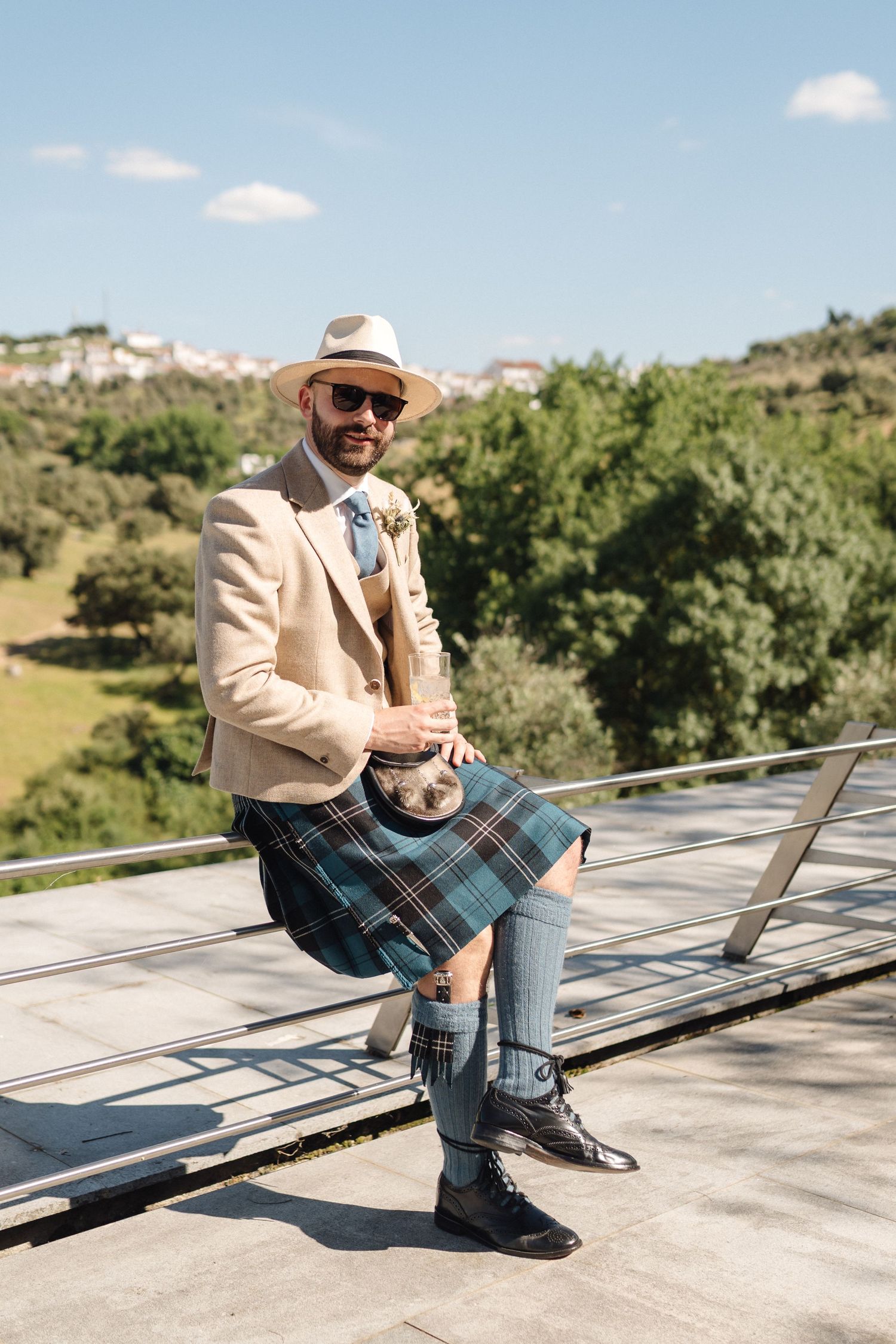 Wedding guest in cream blazer and plaid kilt poses on outdoor terrace.
