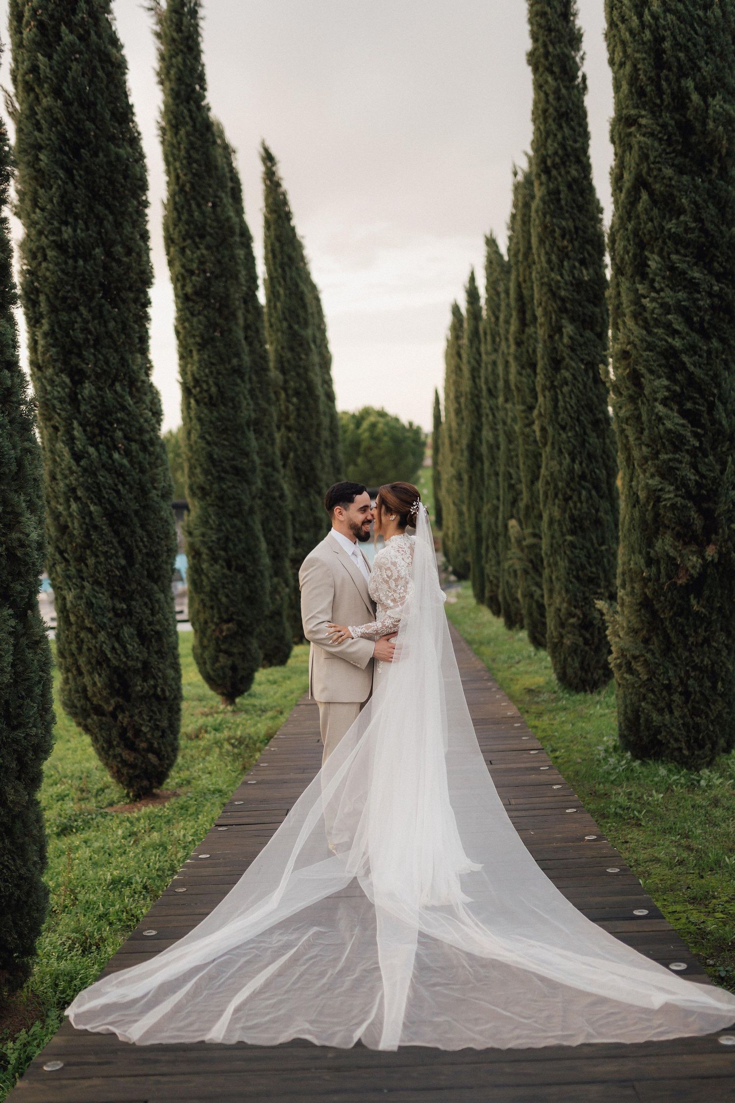 Wedding couple poses romantically along a cypress tree-lined path in an elegant garden setting.