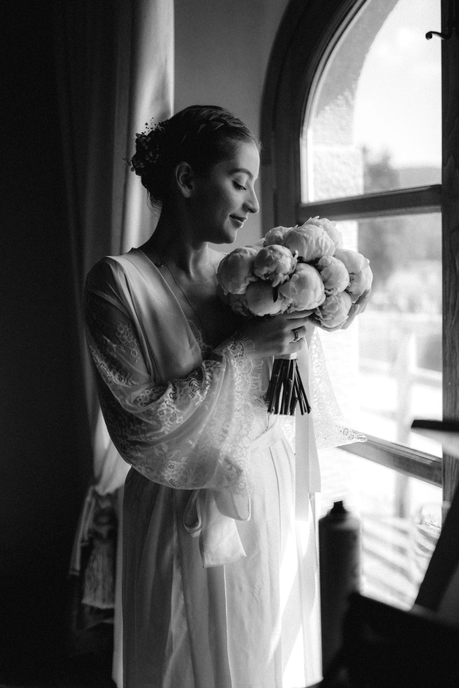 Elegant bride in lace wedding dress holding floral bouquet by arched window in black and white photo.