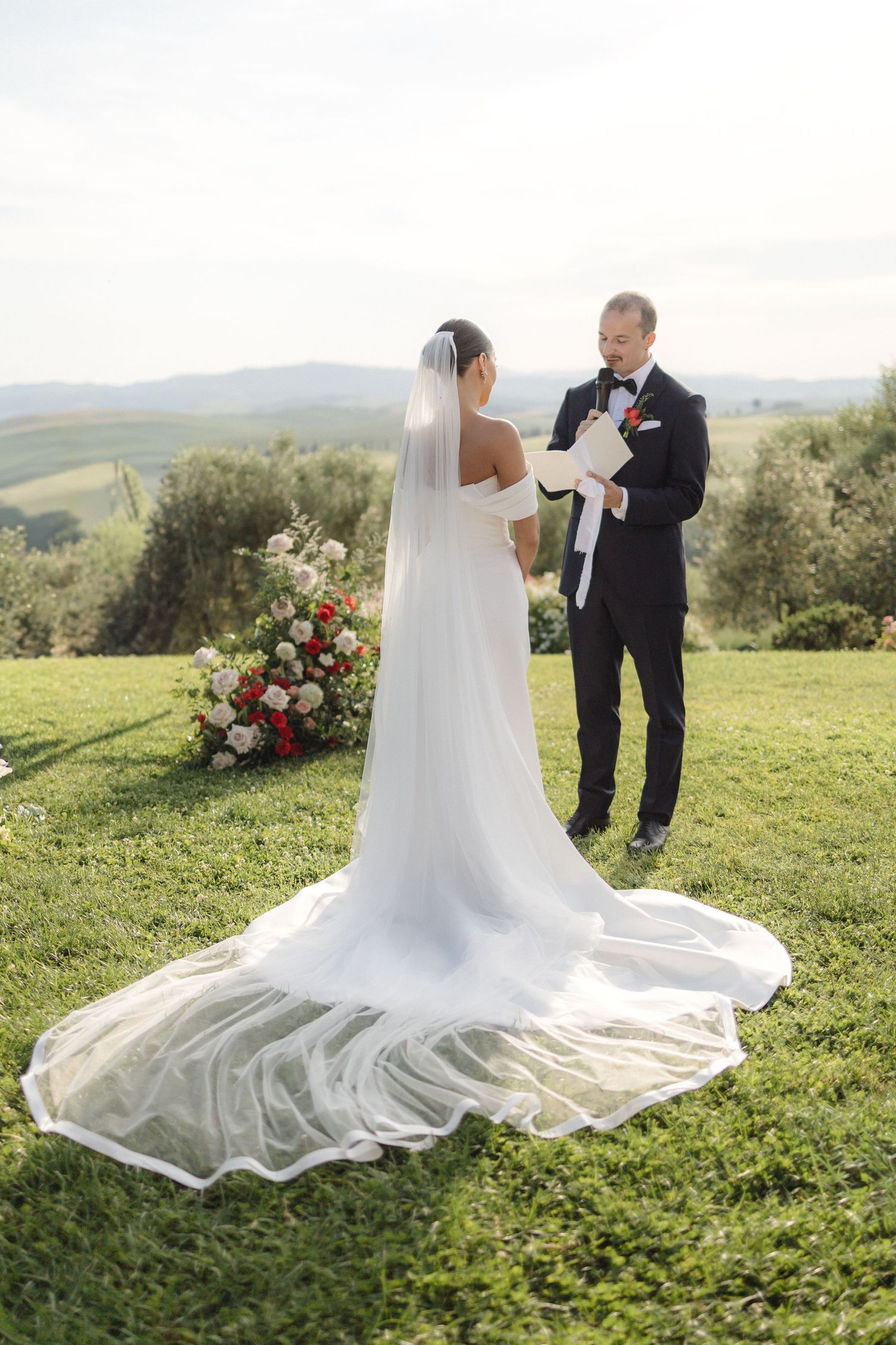 Bride in elegant off-shoulder gown with long cathedral veil exchanges vows with groom in black tuxedo outdoors in Tuscany.