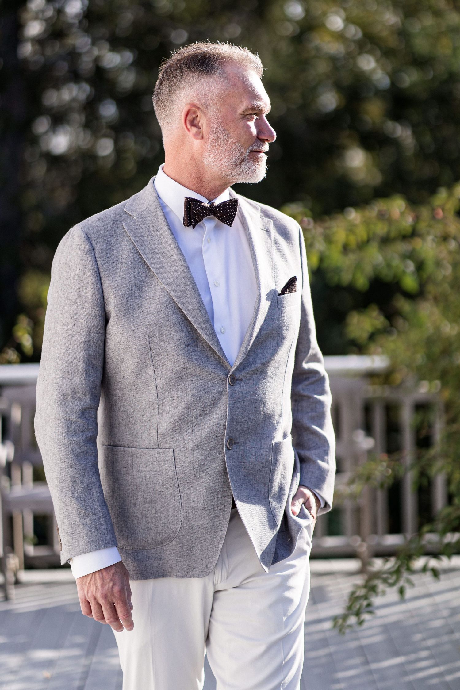 A person wearing a light gray suit jacket and white shirt stands in an outdoor setting.
