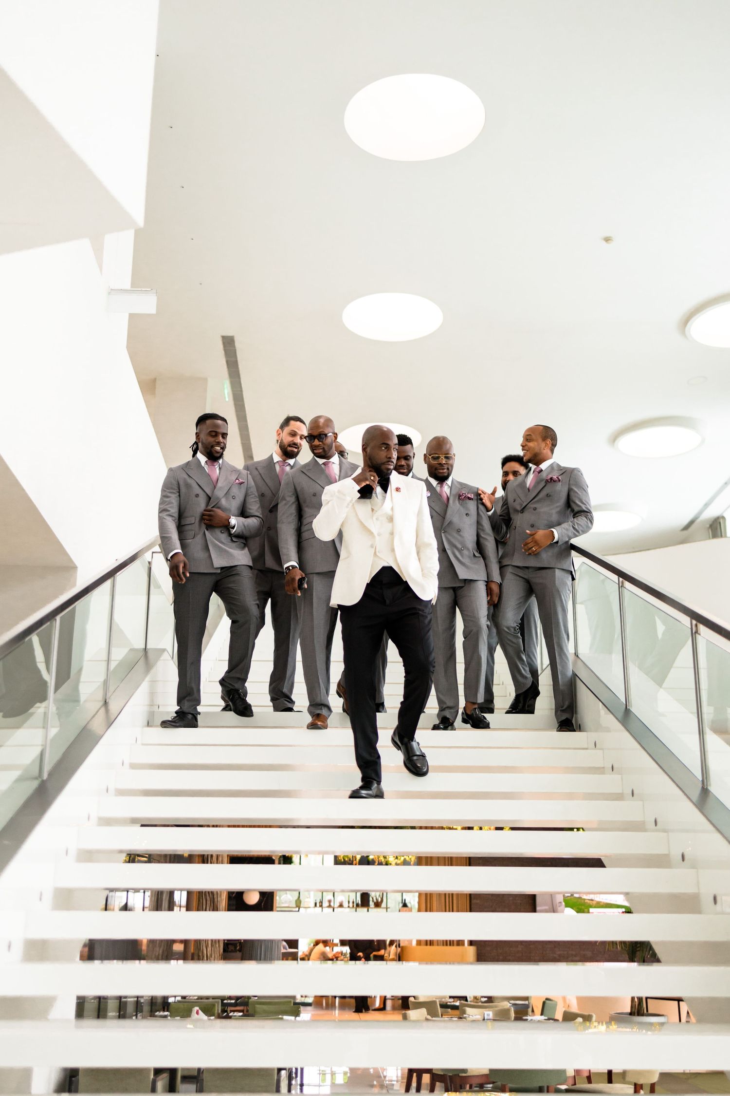 Wedding party members pose on modern white stairs in a contemporary building with circular ceiling lights.