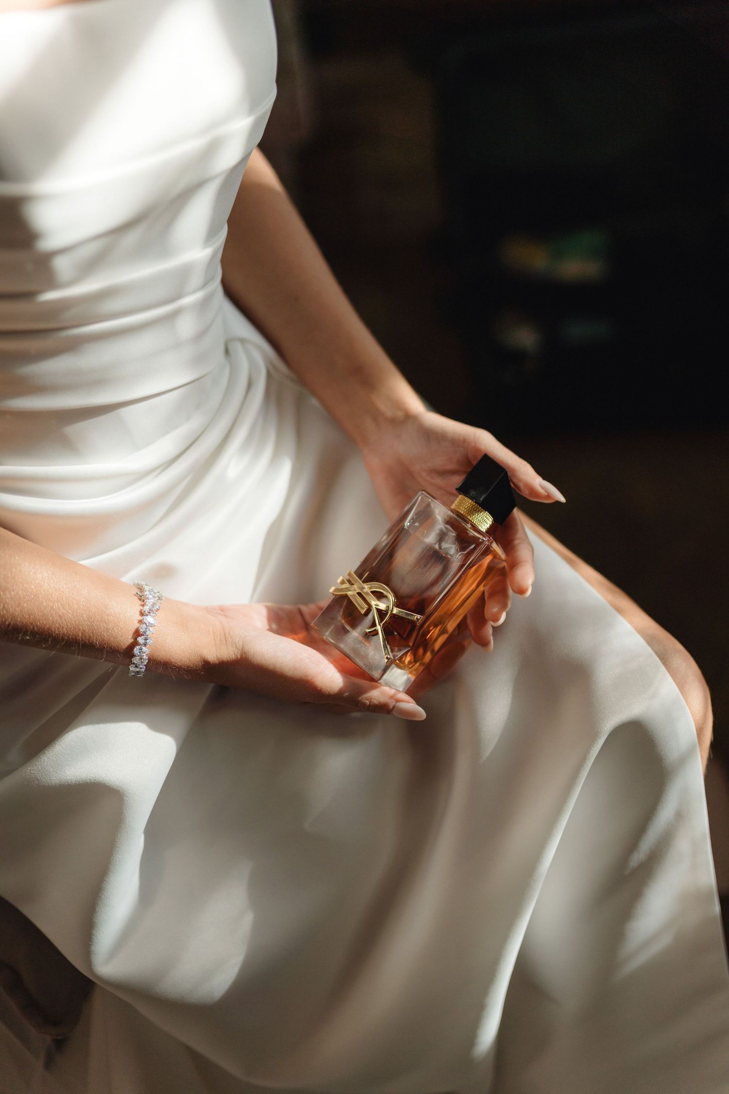 Bride in white satin wedding dress holding a luxury amber perfume bottle with gold accents.