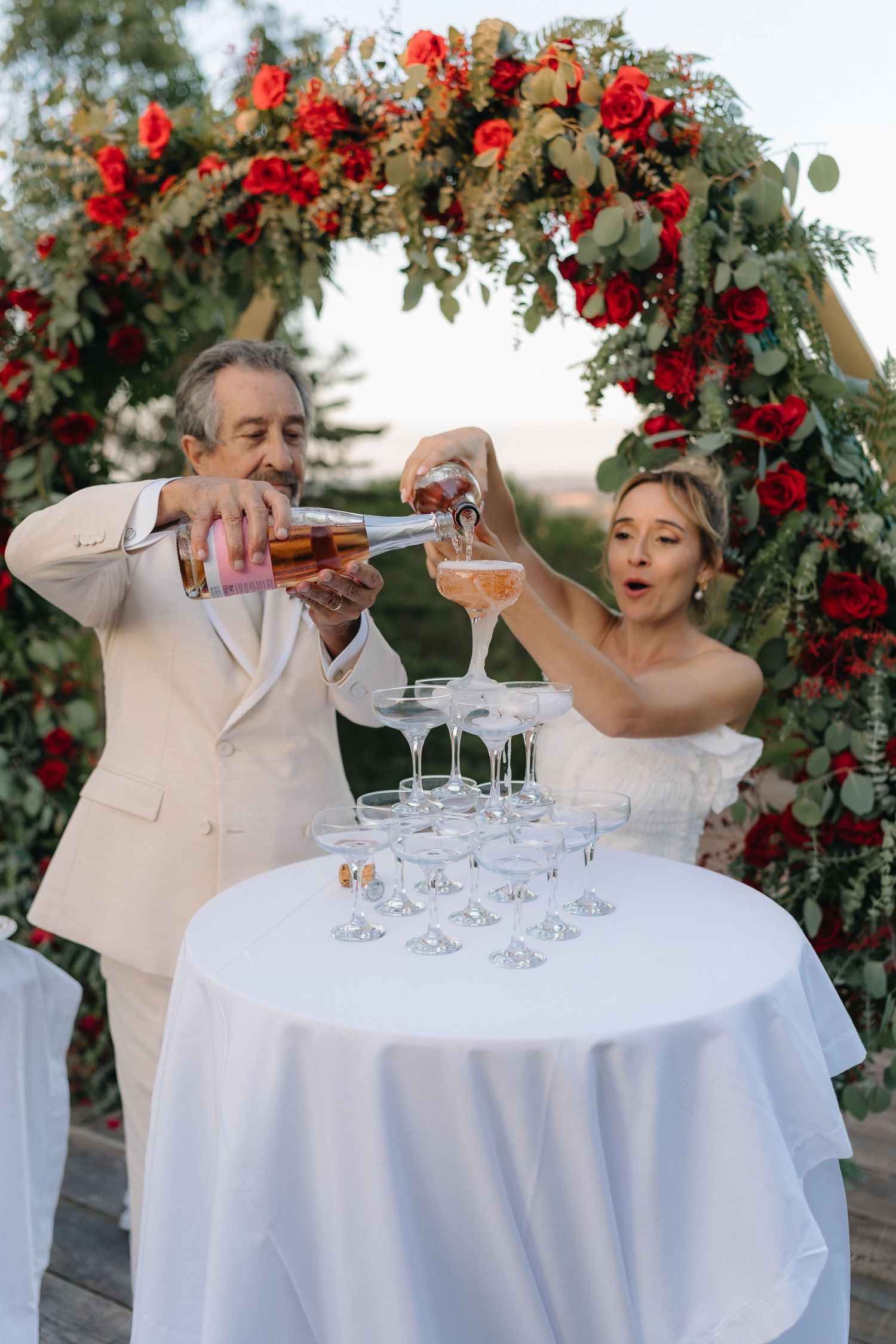 Wedding party creating champagne tower at outdoor reception under floral arch.