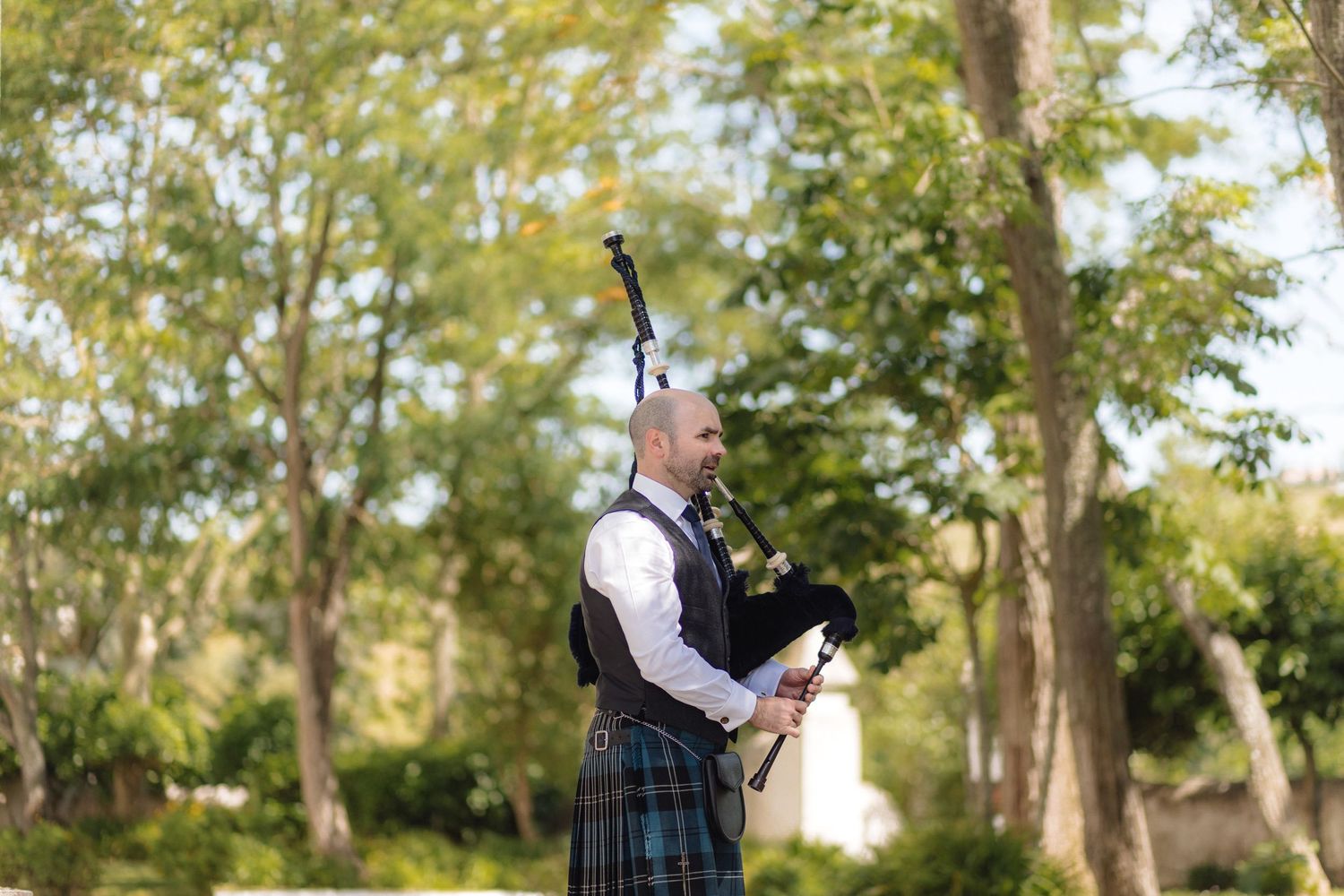 Bagpiper performs outdoors among trees in traditional Scottish attire.