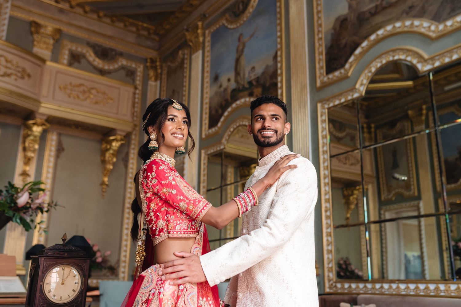 Elegant couple in traditional Indian wedding outfits posing in ornate interior setting.