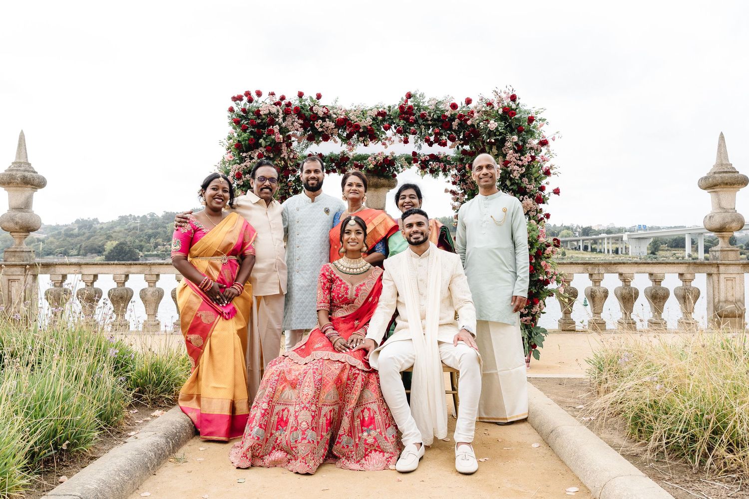 Wedding party poses under floral arch at elegant outdoor venue.