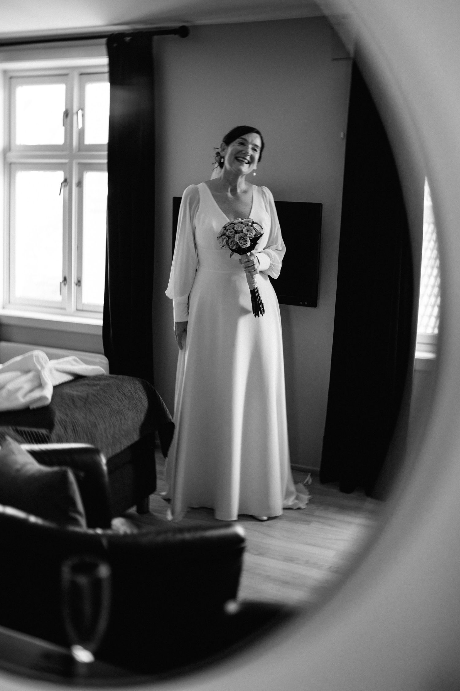A classic black and white photograph of a bride in a long-sleeved wedding dress holding a bouquet in a sunlit room.