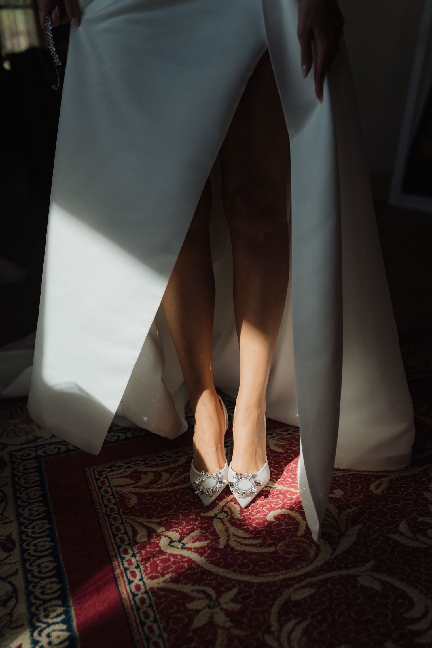 Bride in white wedding gown showing embellished bow heels on ornate red carpet in sunlit room.