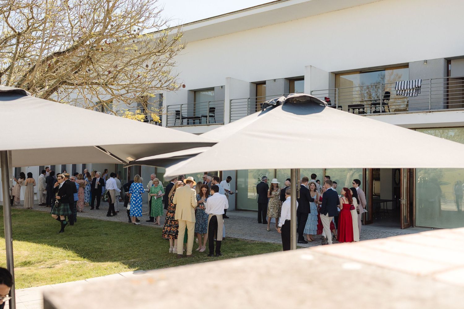 Wedding guests gather under white umbrellas on a patio outside a modern white building.